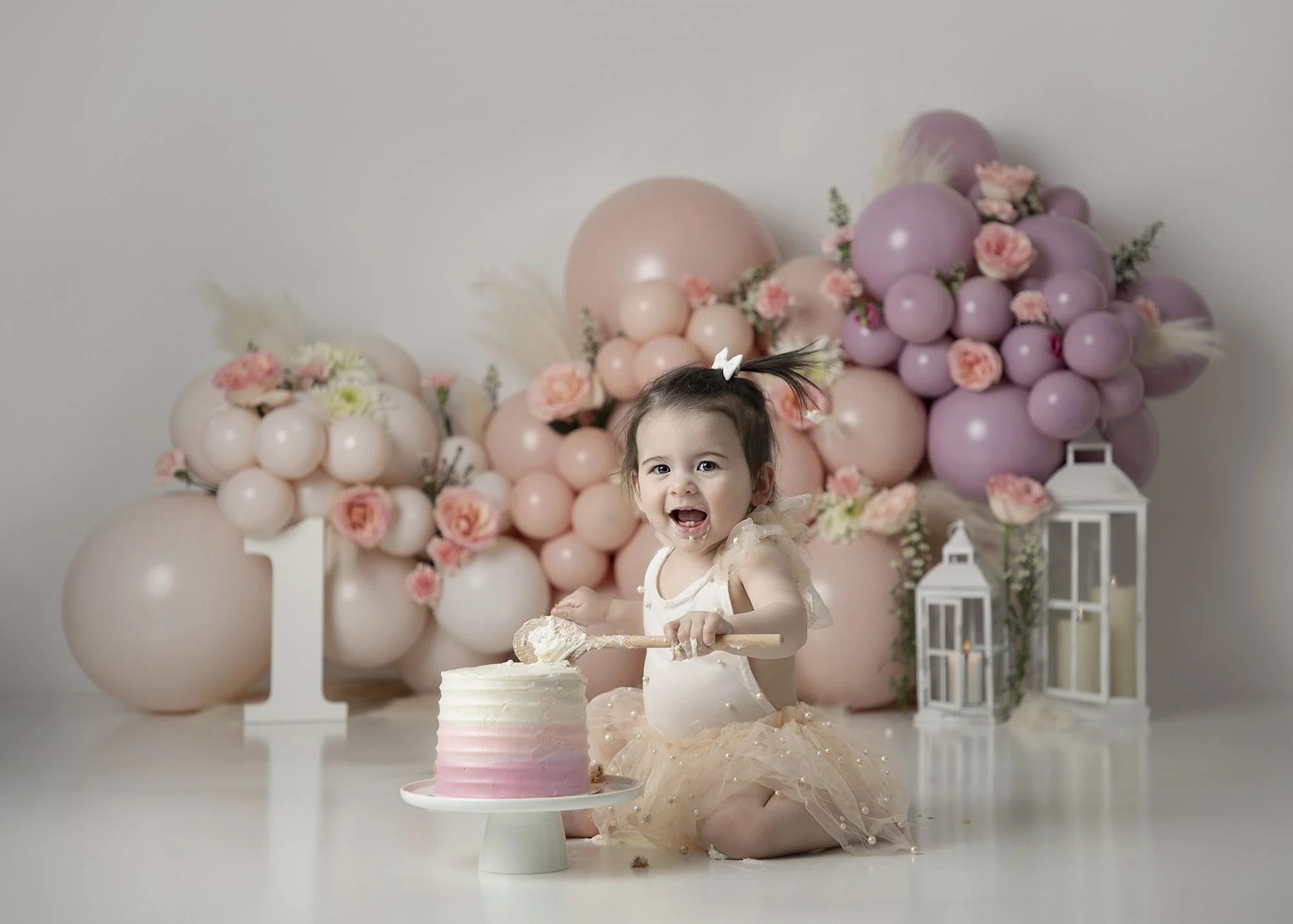 Young girl with a bow in her hair celebrating a birthday with a pink and white cake, surrounded by pink, purple, and white balloons, flowers, and lanterns.