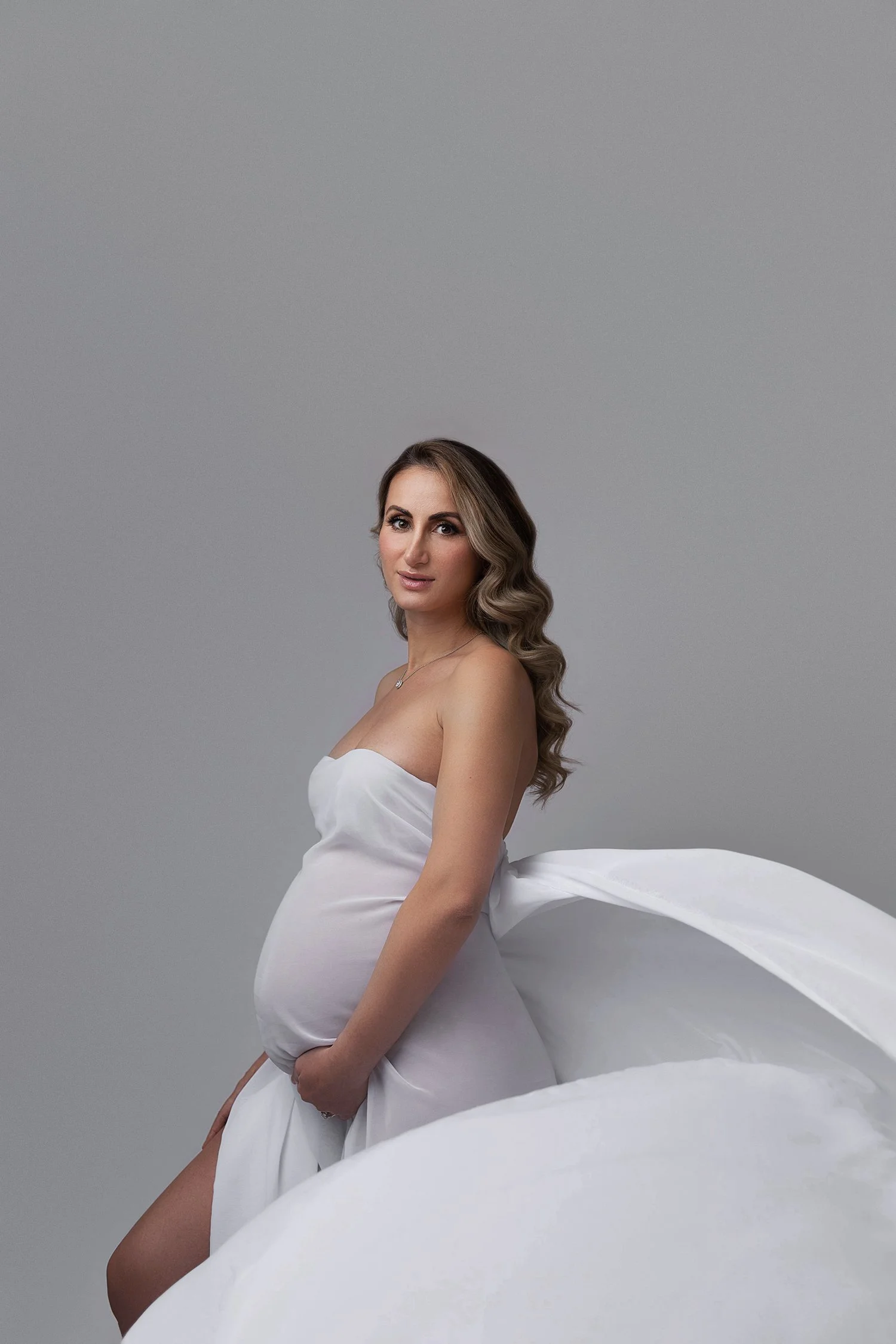 A pregnant woman with long wavy hair in a strapless white dress, sitting against a plain gray background, looking at the camera.