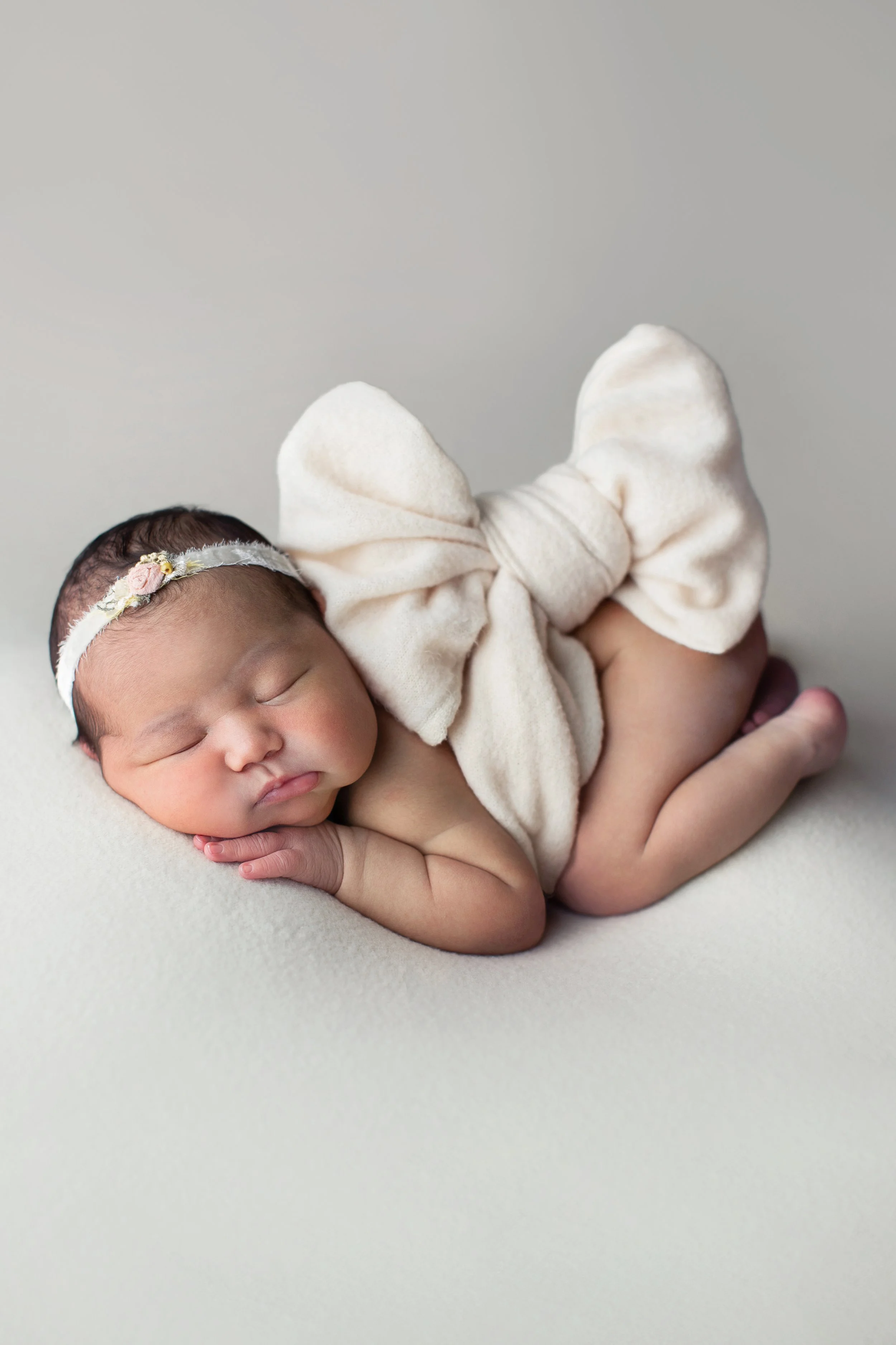 Close-up of a sleeping newborn baby with a flower headband and large cream bow on their head, lying on a soft white surface.