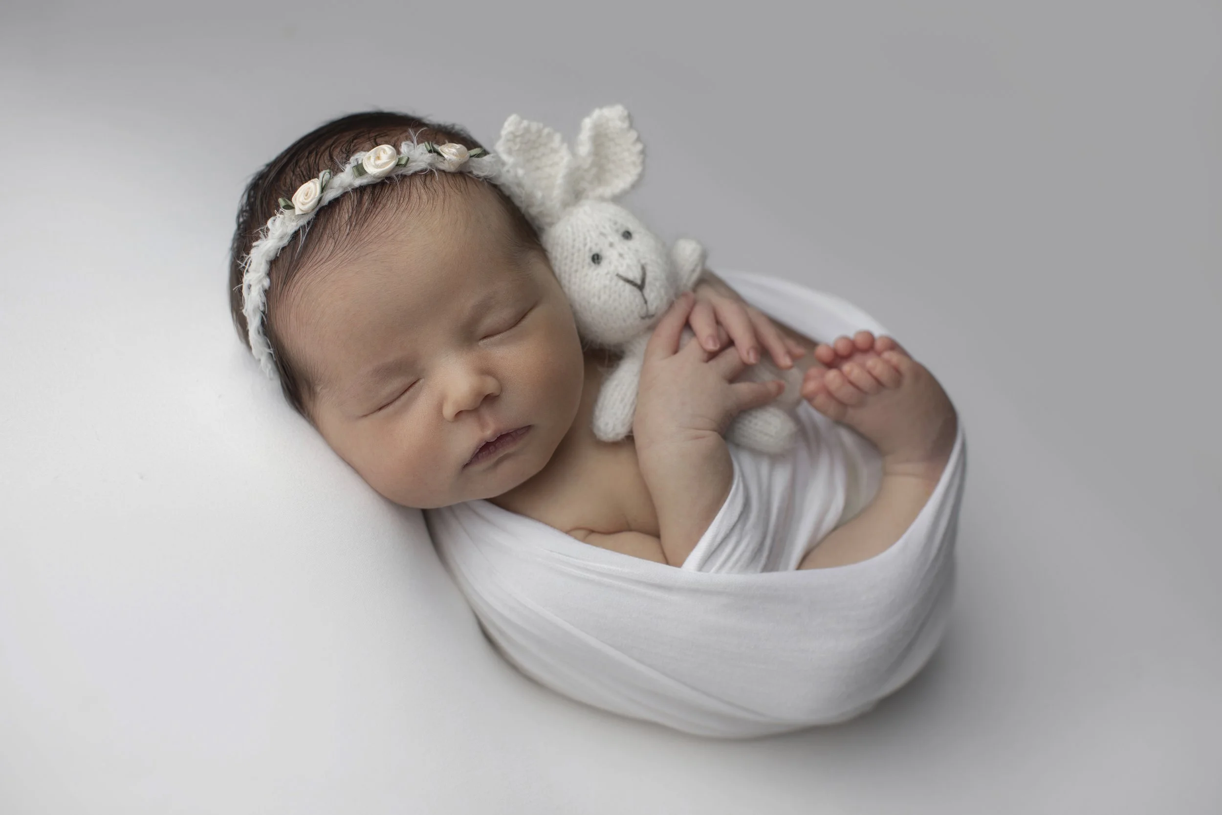 A sleeping newborn baby wrapped in white cloth and holding a small, white, knitted teddy bear, wearing a floral headband.
