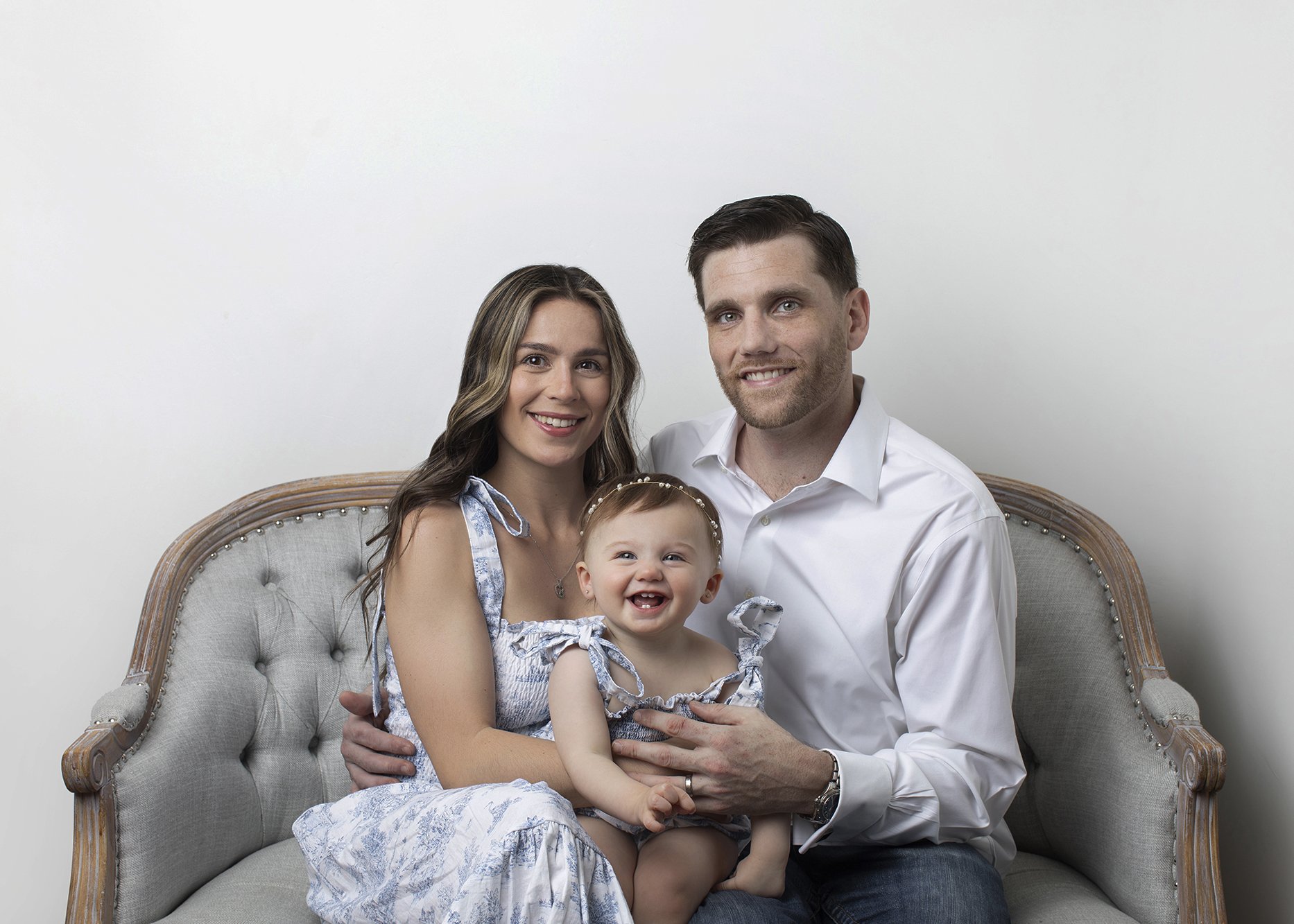 A happy family of four sitting on a vintage-style chair against a plain white wall. The mother has long, wavy brown hair, and the father has short dark hair and a beard. The little girl, sitting on the mother's lap, is smiling and has short brown hai