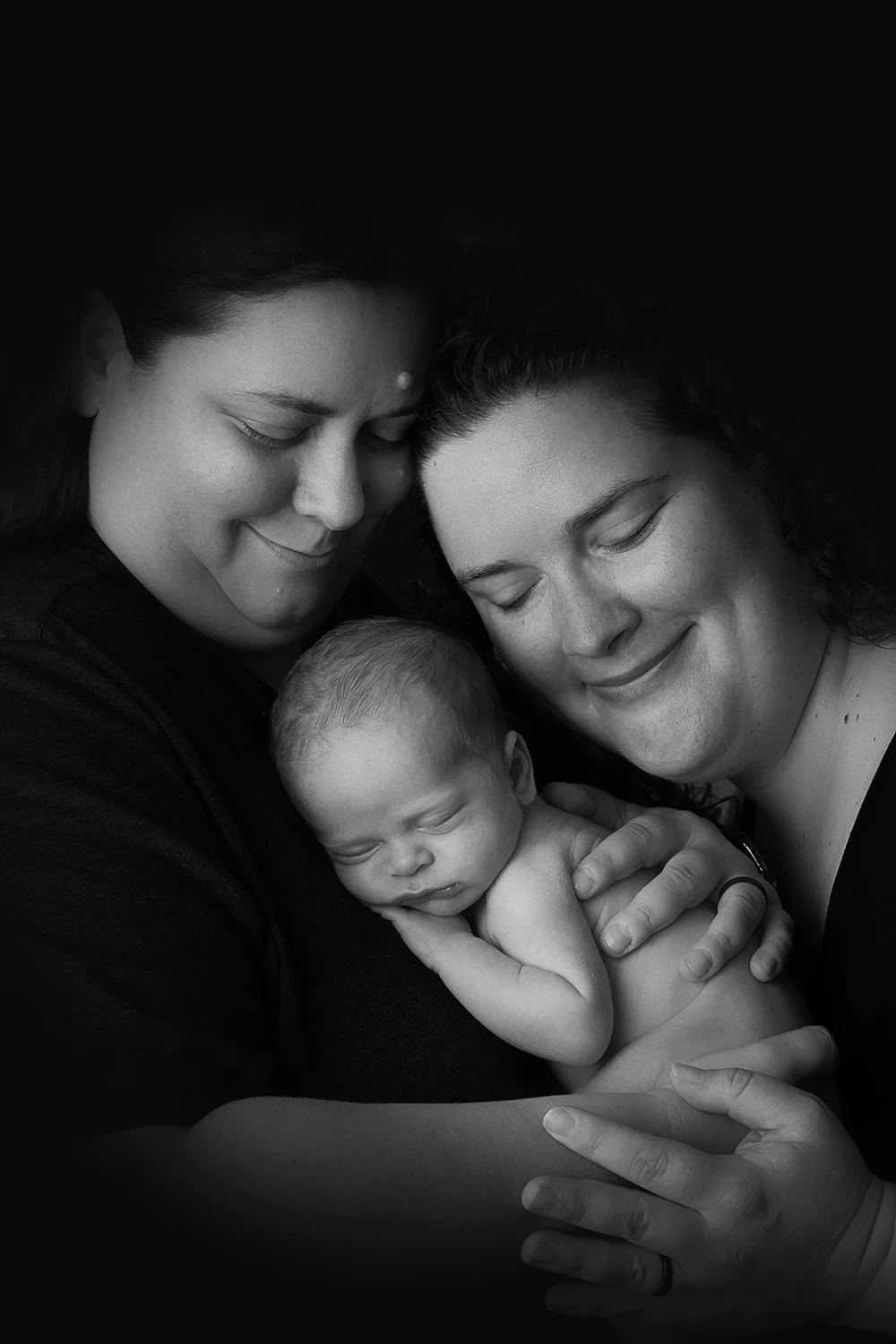 Black and white photo of two women holding a sleeping baby, all with gentle smiles, close together in an intimate pose.