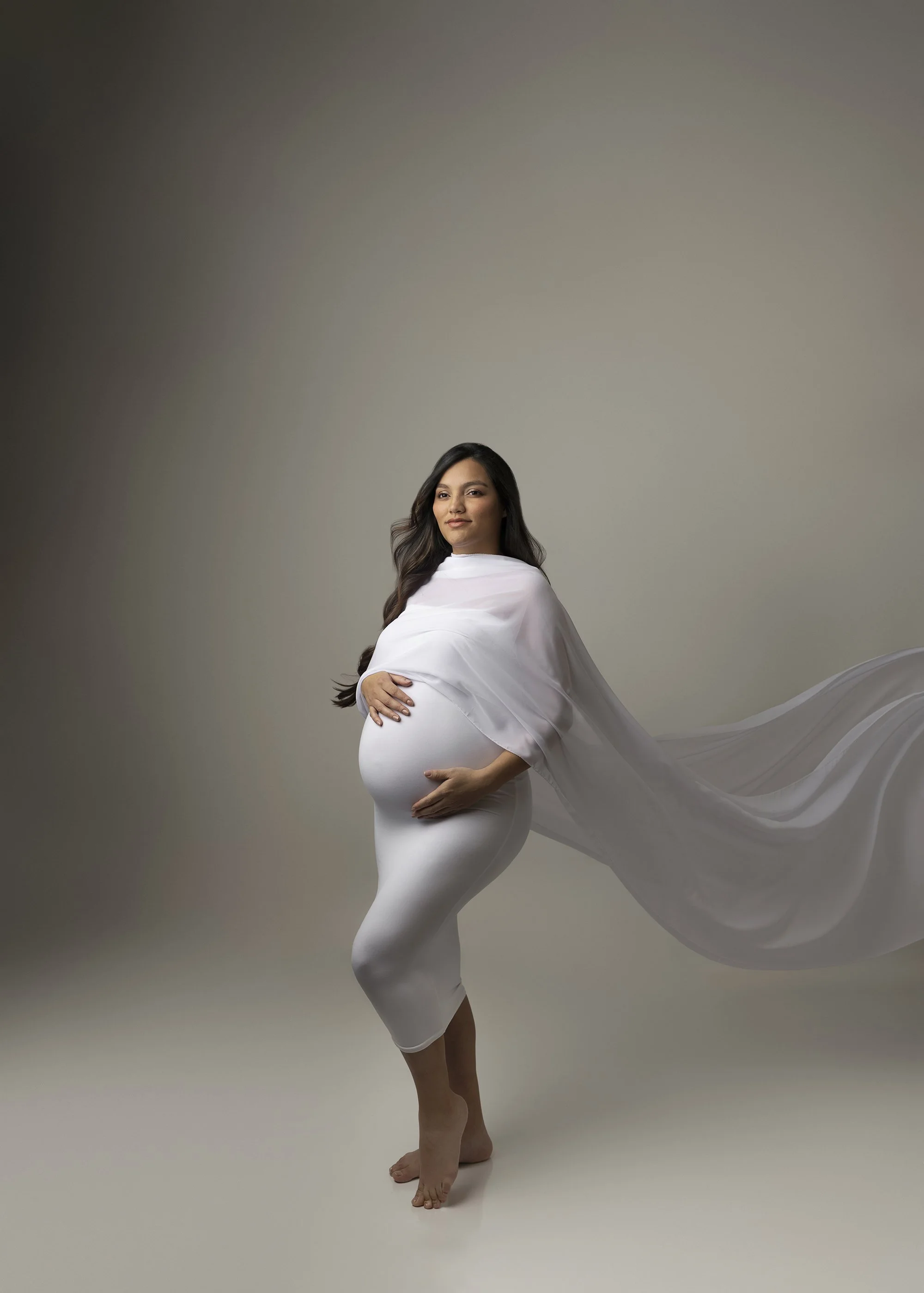 A pregnant woman in a white dress and flowing cape poses on a plain background.