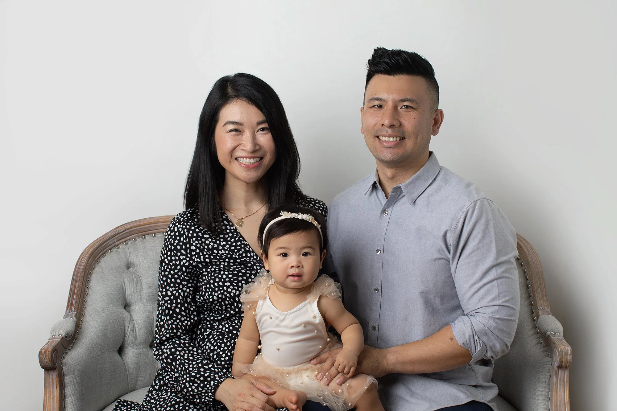 A family of three, including a woman, man, and young girl, posing together for a portrait against a plain white background. The woman has black hair, the man has short black hair, and the girl is wearing a light-colored dress with a tulle skirt and a
