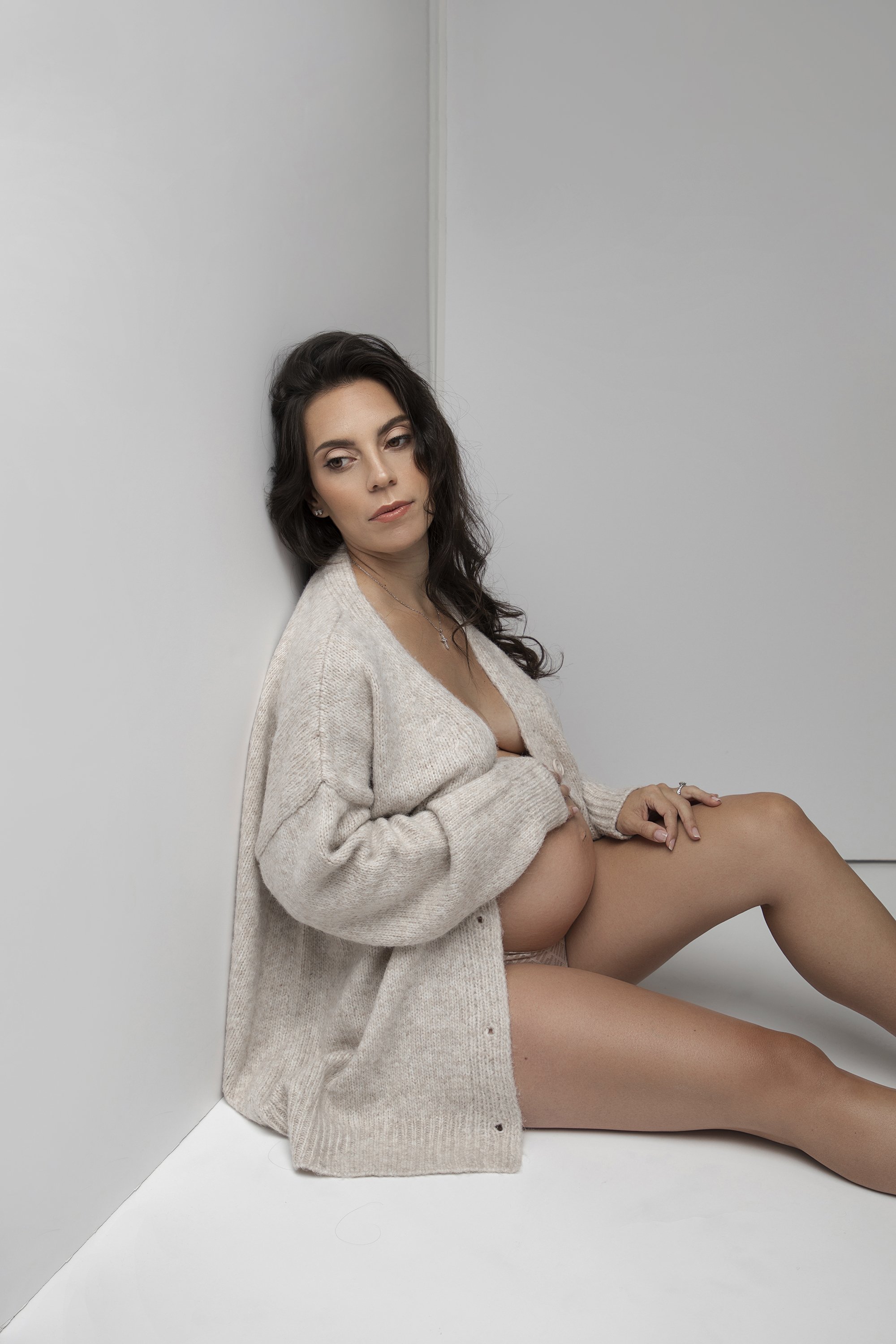 A woman with dark hair and light skin, sitting against a corner with white walls, wearing a beige cardigan, holding her legs, with a calm expression.