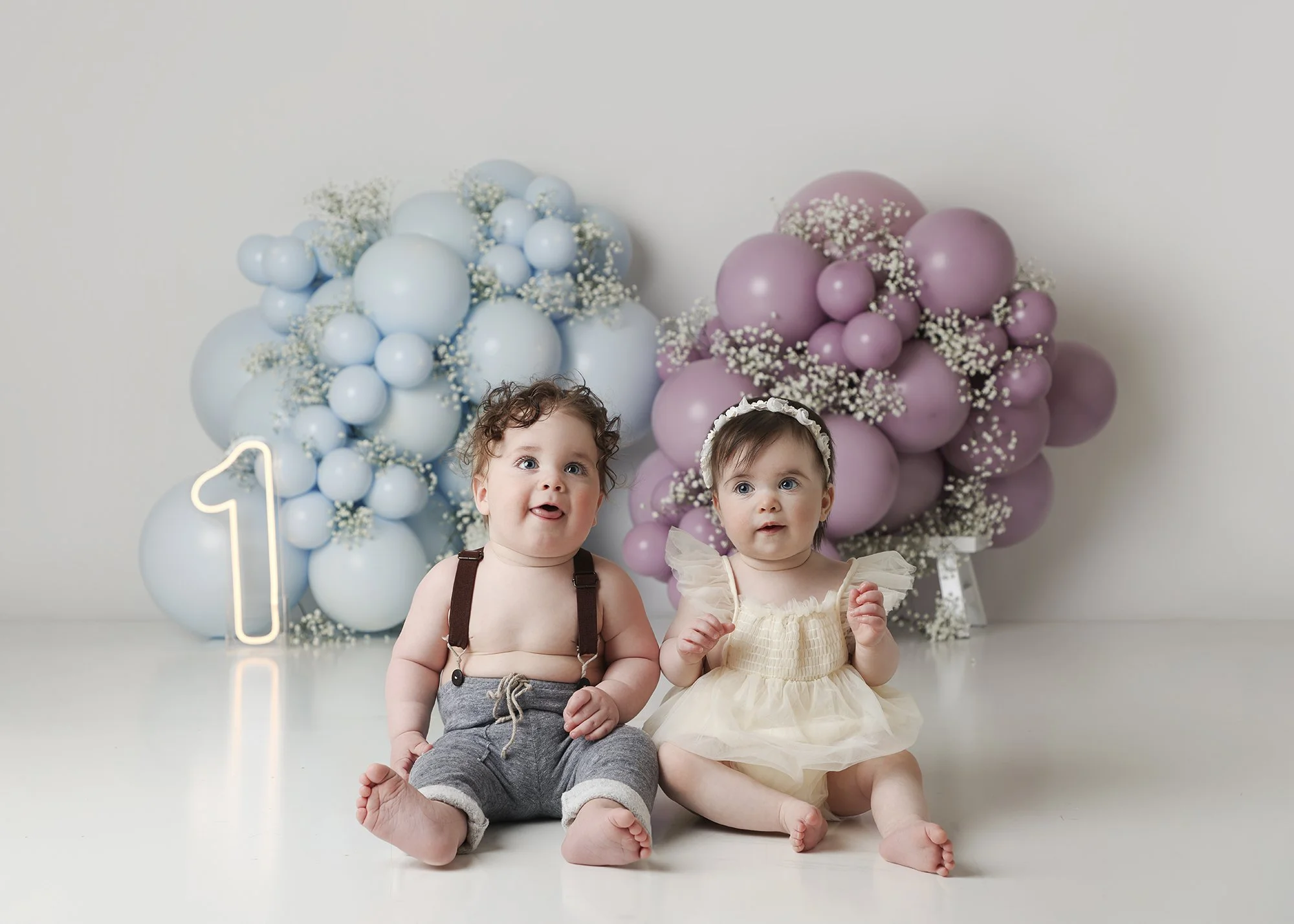 Two young children sitting on the floor in front of pastel-colored balloon decorations with a number one lit-up sign, celebrating a first birthday.