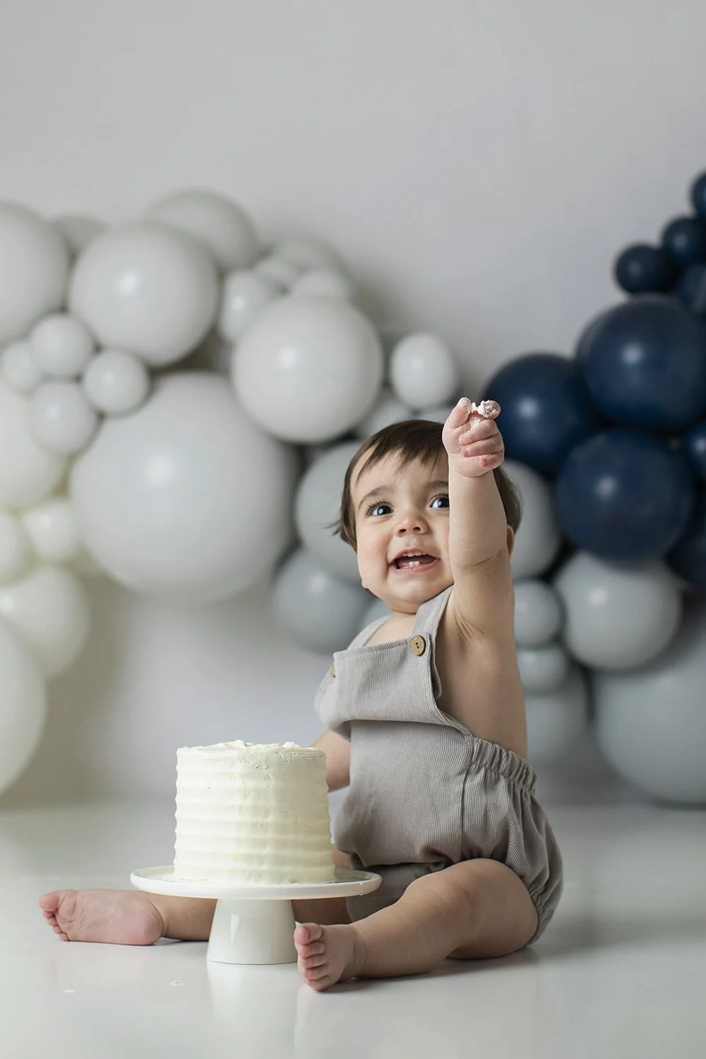 A happy baby sitting in front of a small white cake on a cake stand, reaching upward with one hand, with gray and navy blue balloon decorations in the background.