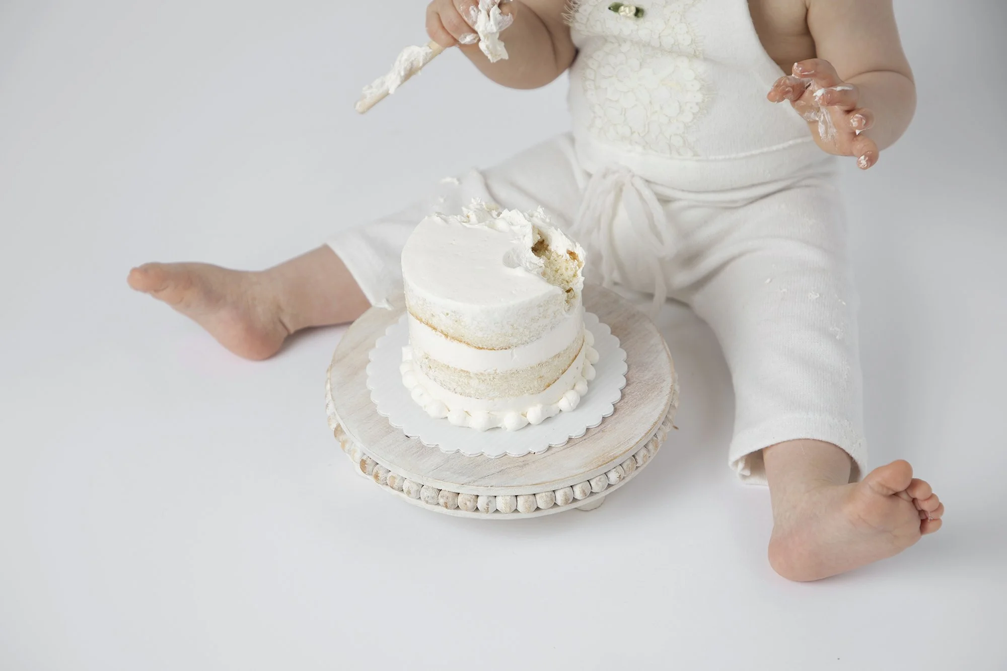 A young child wearing a white outfit is sitting on a surface and holding a piece of a partially eaten white cake on a wooden cake stand in front of them.
