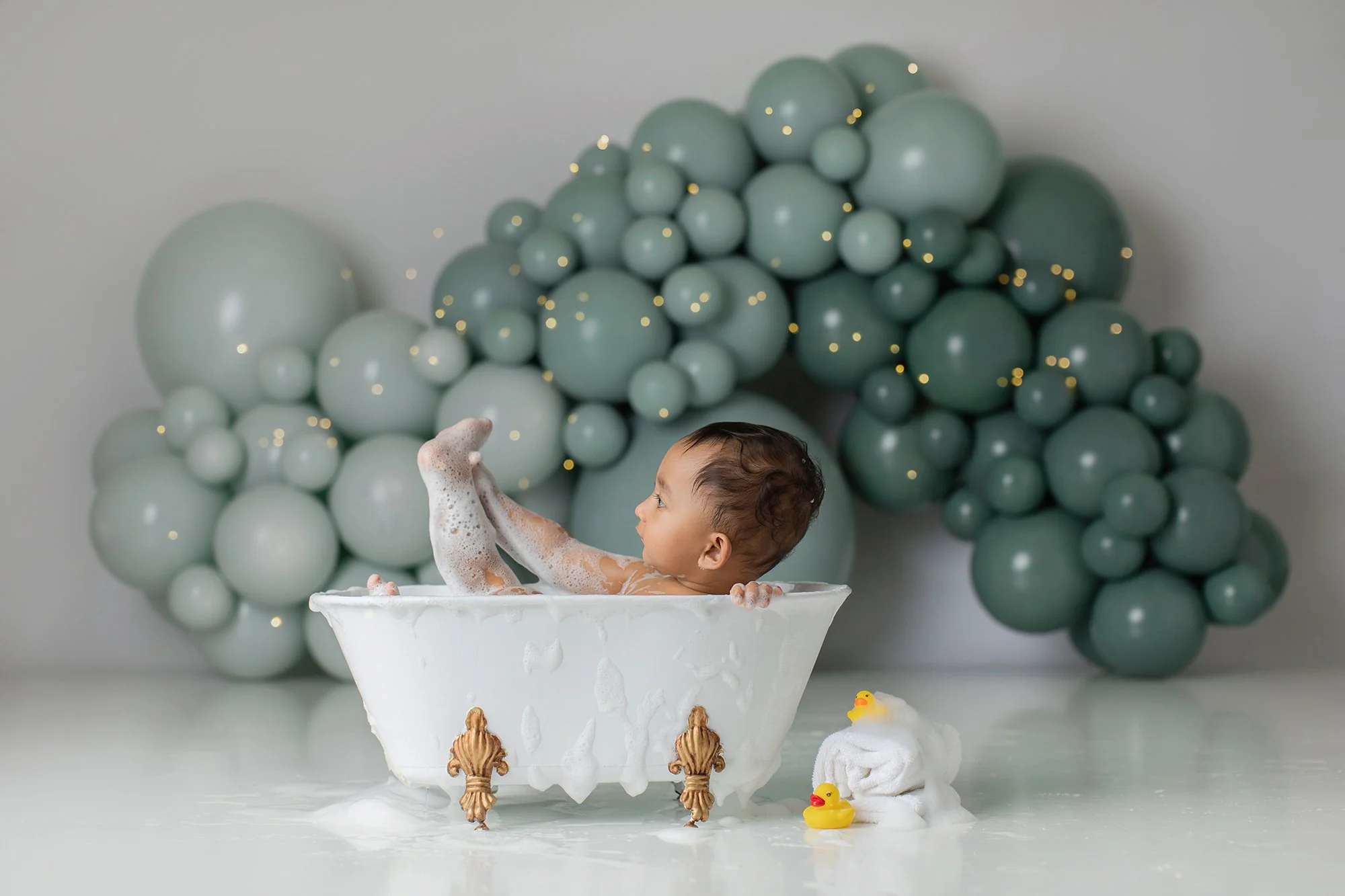 A young child taking a bubble bath in a vintage clawfoot bathtub with a backdrop of greenish-gray balloons. Two rubber duck toys are nearby, and the child is playing with foam bubbles.