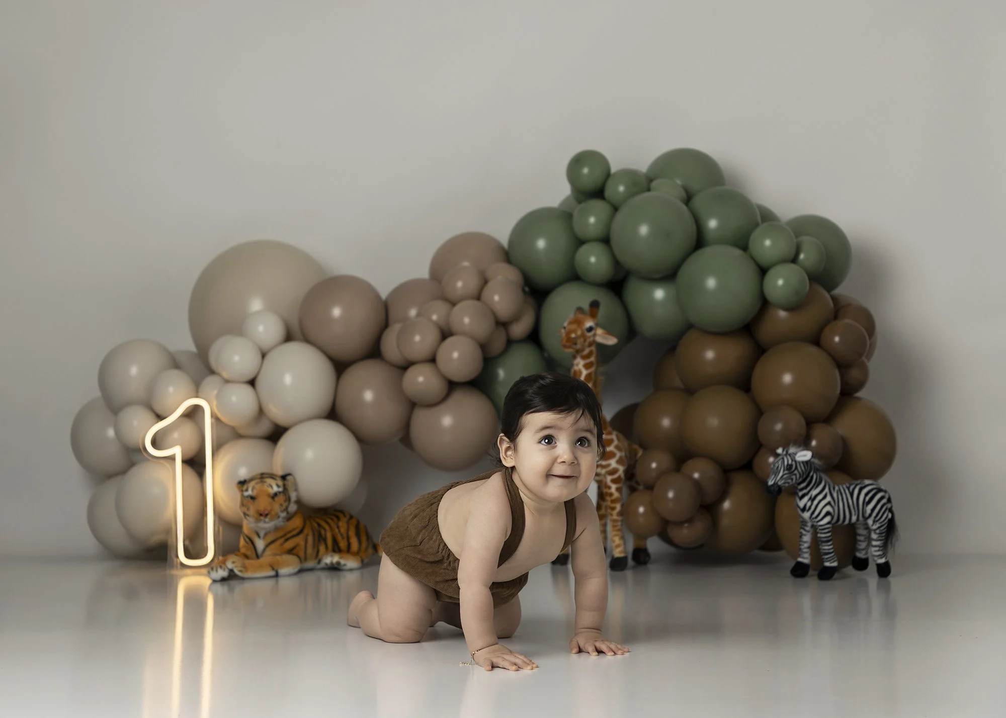 Young boy crawling on the floor in front of a backdrop of neutral-colored balloons, tiger and zebra plush toys, and a decorative letter 'N' light.