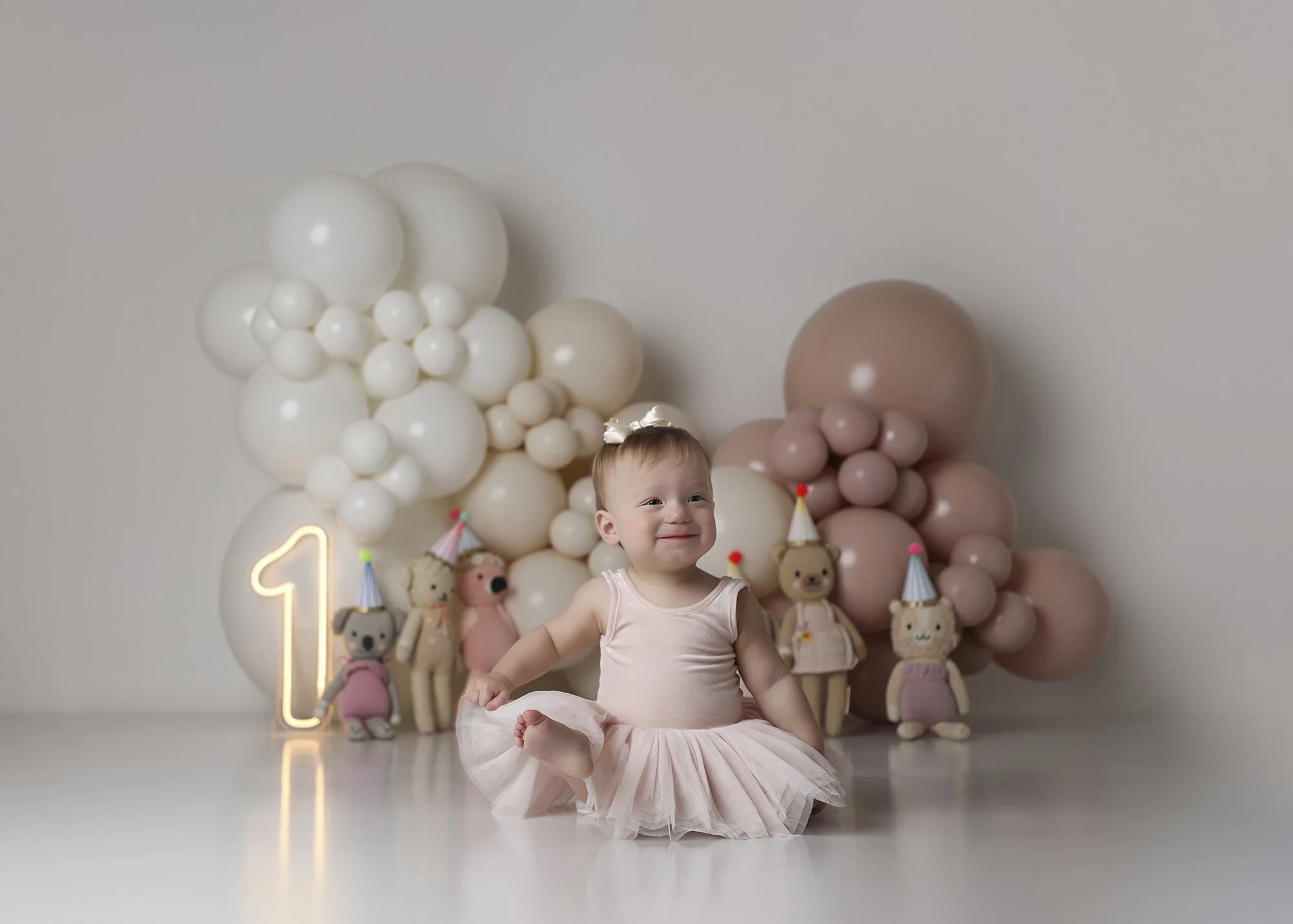 A smiling young girl wearing a pink dress and tutu, sitting on the floor in front of a balloon backdrop with teddy bear decorations.