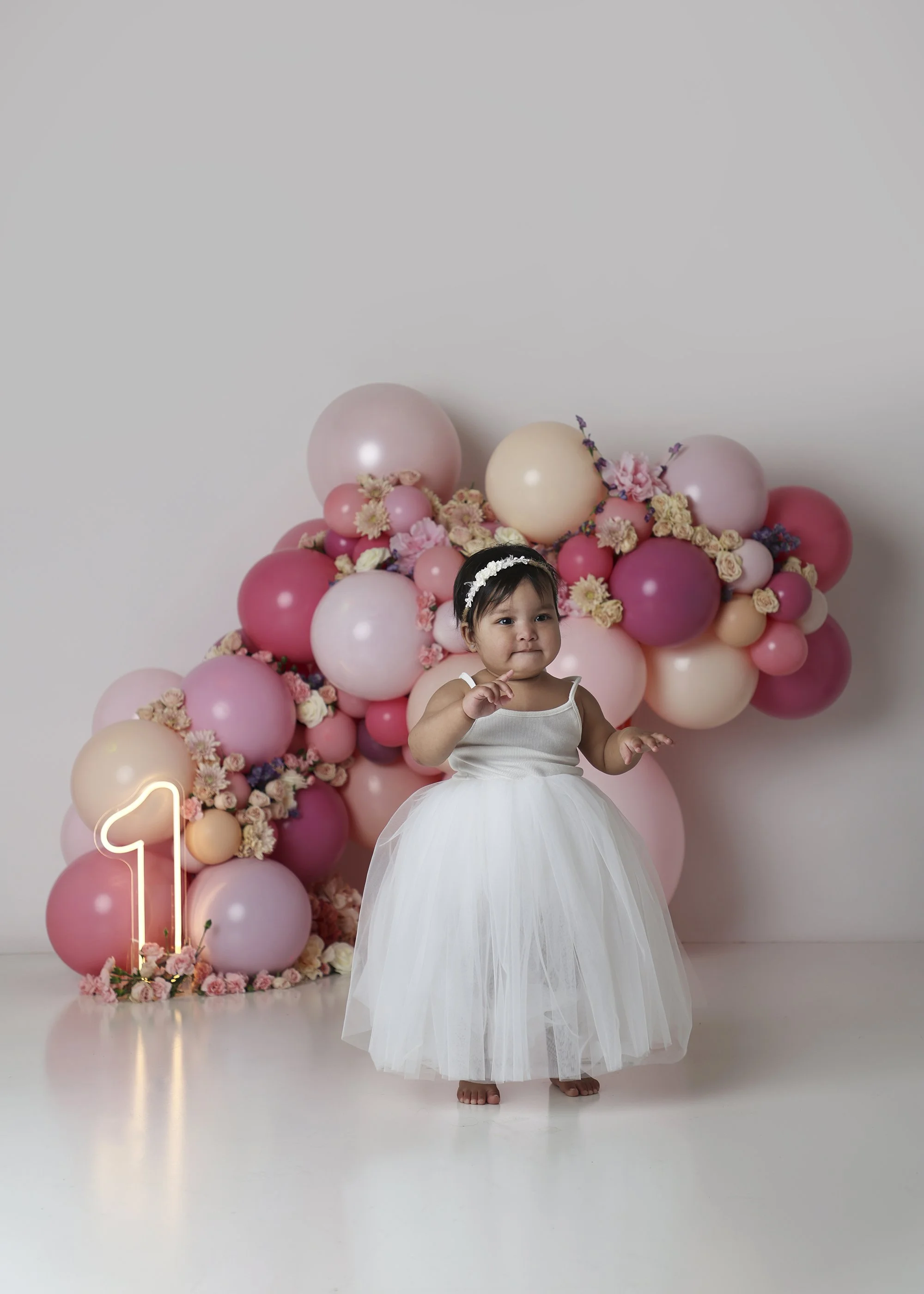 A young girl wearing a white dress and a floral headband stands in front of a pink balloon and flower backdrop, with a neon number one sign on the floor, celebrating her first birthday.