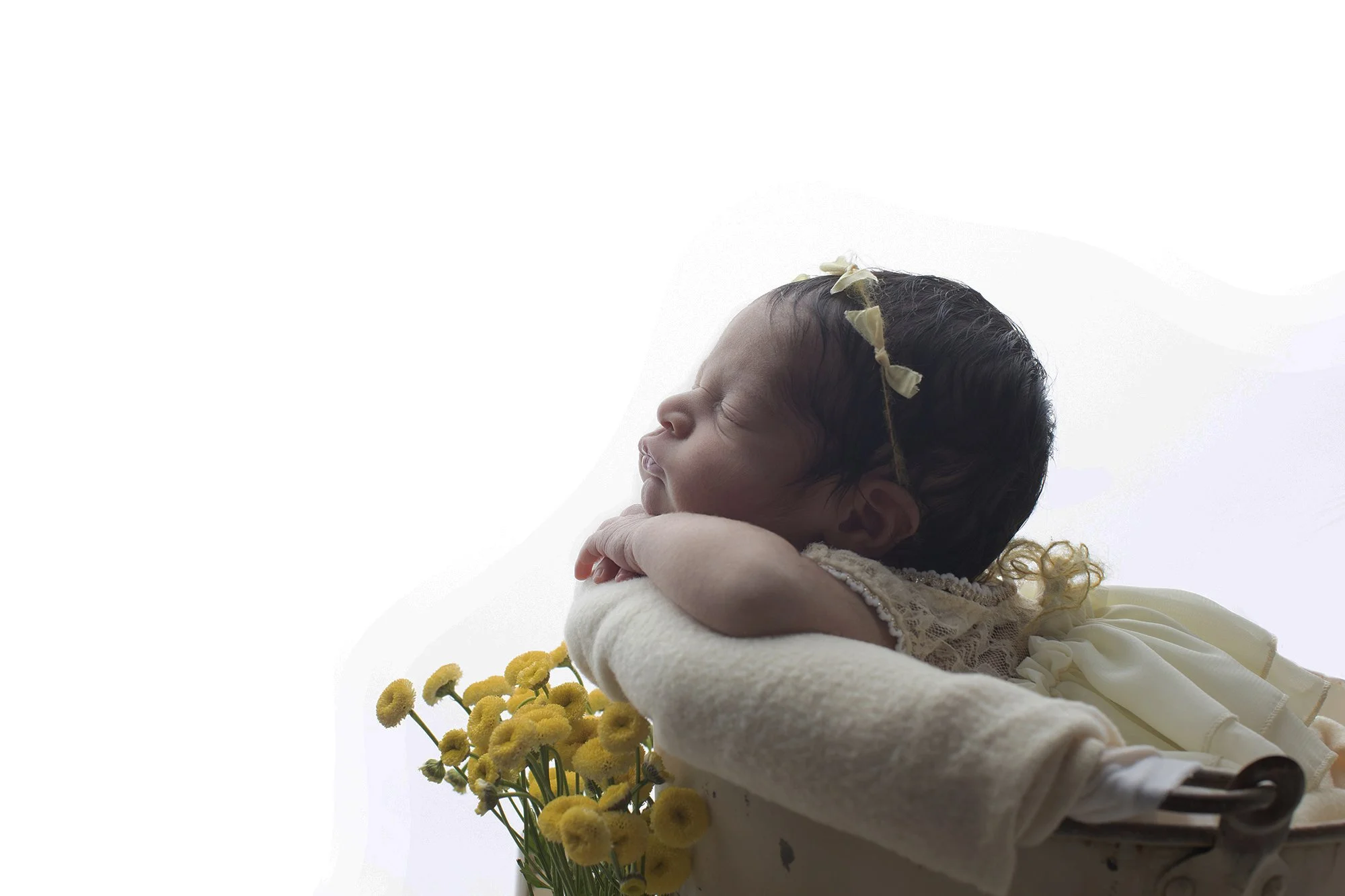 A newborn baby with dark hair and a flower crown, dressed in light-colored clothing, resting on a soft surface with yellow flowers nearby.