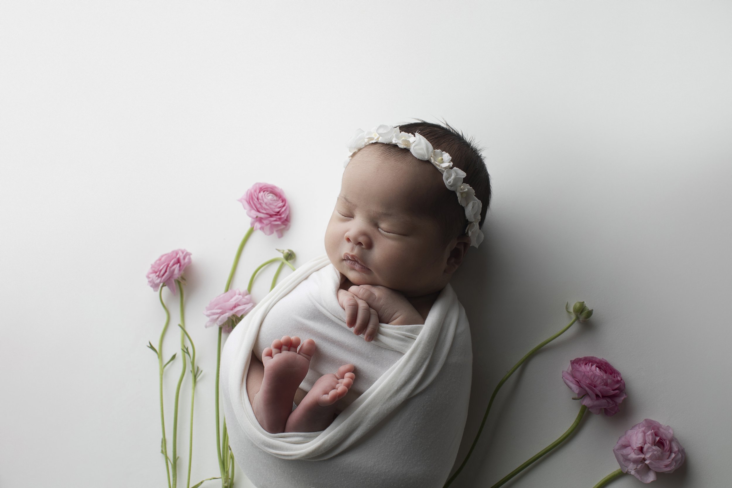 A sleeping baby wearing a white headband with white flowers, swaddled in a white blanket, lying on a white surface surrounded by pink ranunculus flowers.