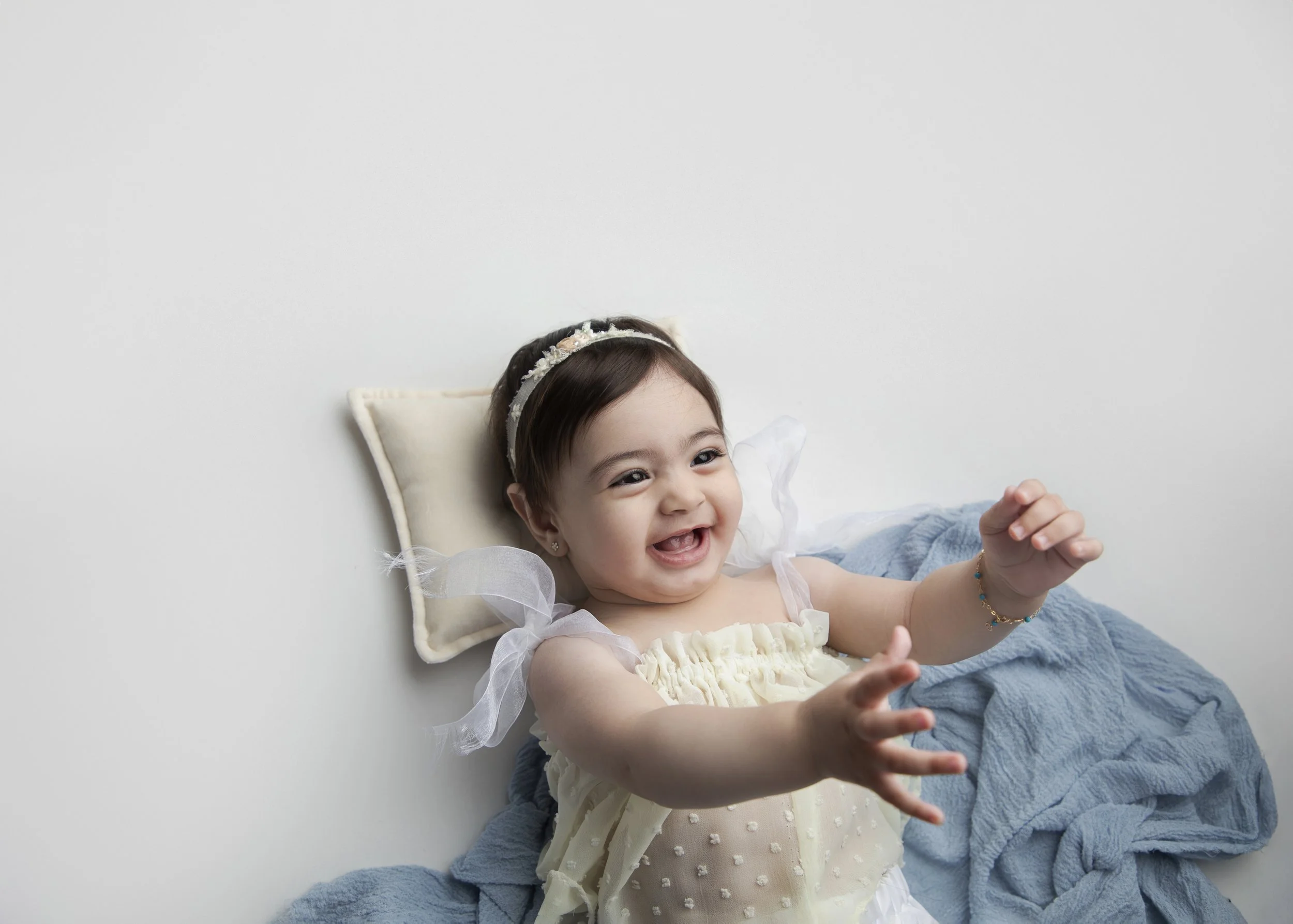 A young girl lying on a bed with a pillow behind her head, smiling and reaching out with one hand, wearing a dress with puffed sleeves and a headband, with a blue blanket partially covering her.