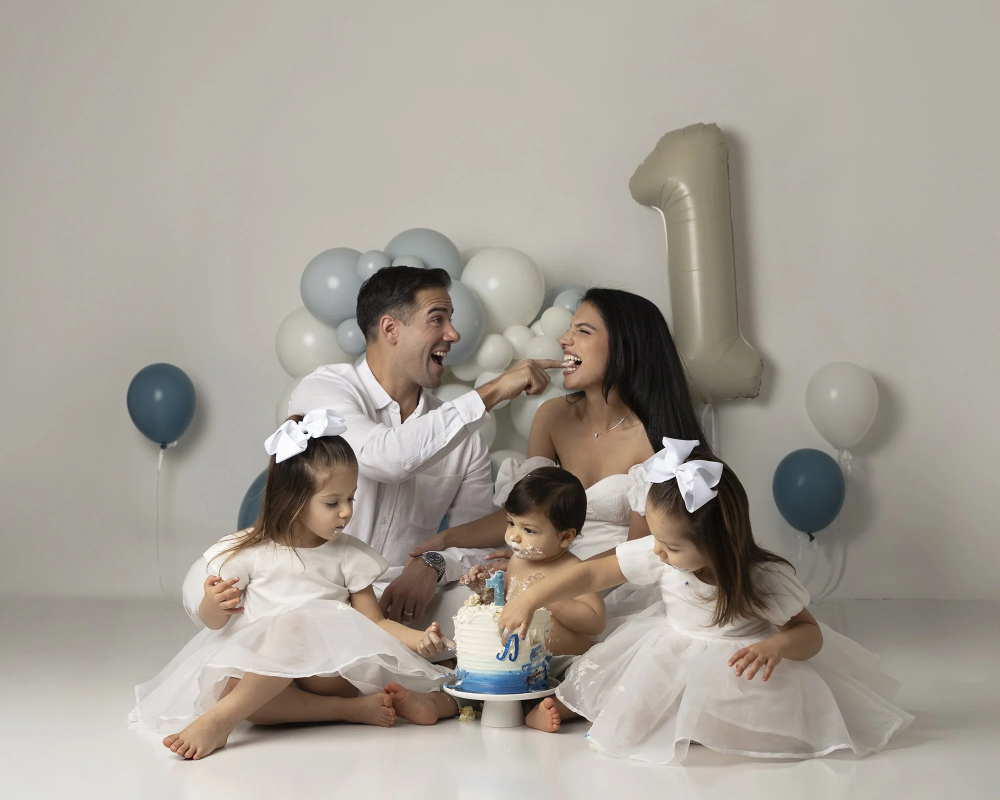 A family celebrating a child's first birthday with a cake, surrounded by balloons and a large number 1 balloon, in a room with a white wall.