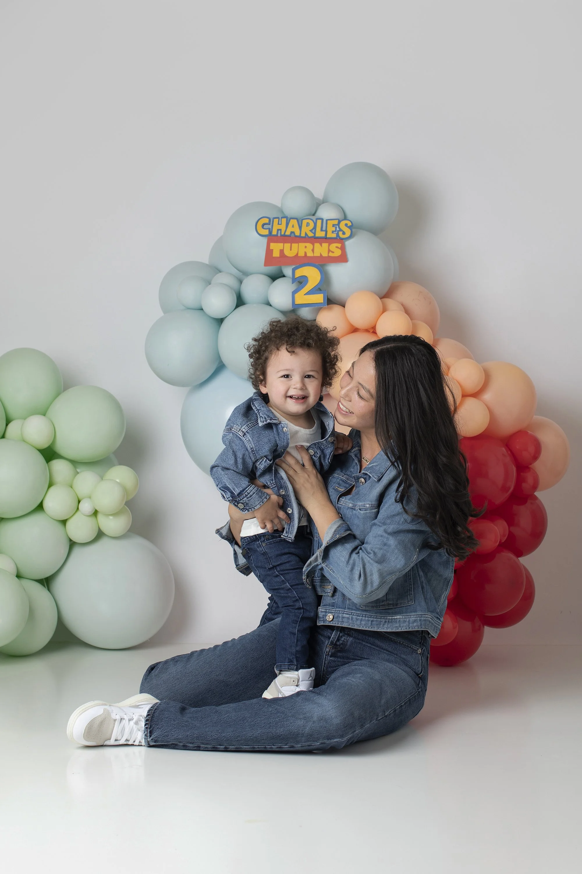 A woman sitting on the floor holding a young child, both smiling, with colorful balloons and a sign that reads "Charles Turns 2" in the background.