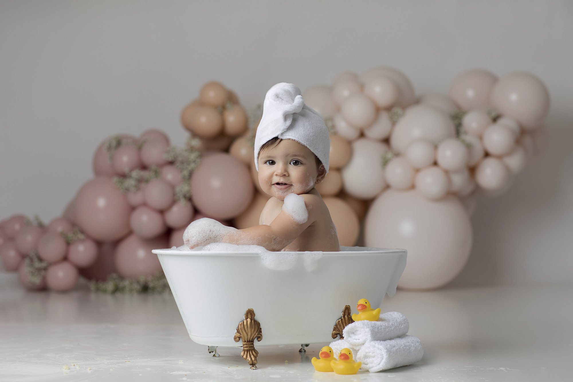 A young child with a white towel wrapped around their head sits in a small white bathtub filled with soap bubbles, smiling and looking at the camera. The background features an arrangement of pink and beige balloons and some greenery. In front of the
