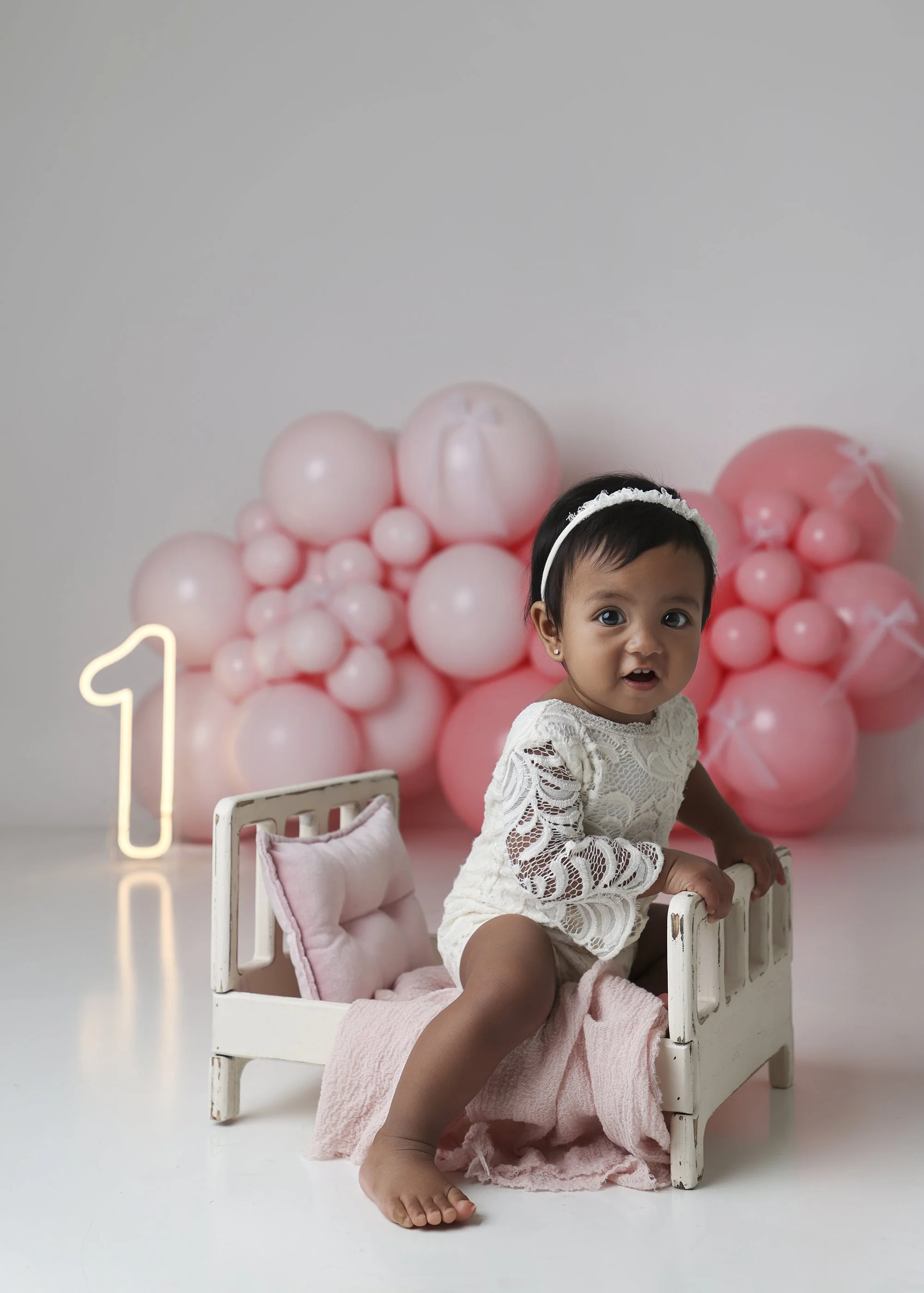 A young girl with short black hair sits on a small white wooden bed, wearing a white lace dress and a white headband, with a pink blanket draped over her legs. Behind her, there is a large backdrop of pink balloons arranged in clusters, with a neon n