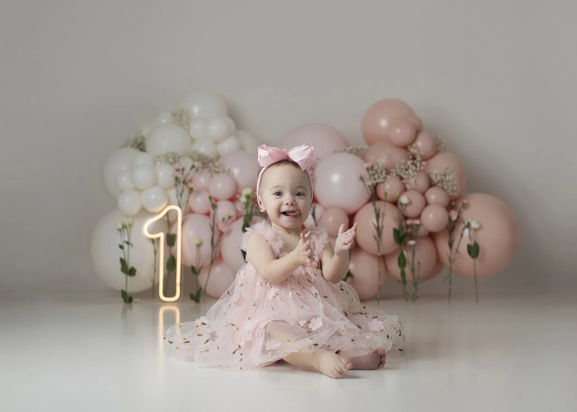 A young child sitting on the floor in front of a backdrop of pink and white balloons, wearing a pink tutu dress with floral details and a large pink bow headband, with a neon number one sign to the side.