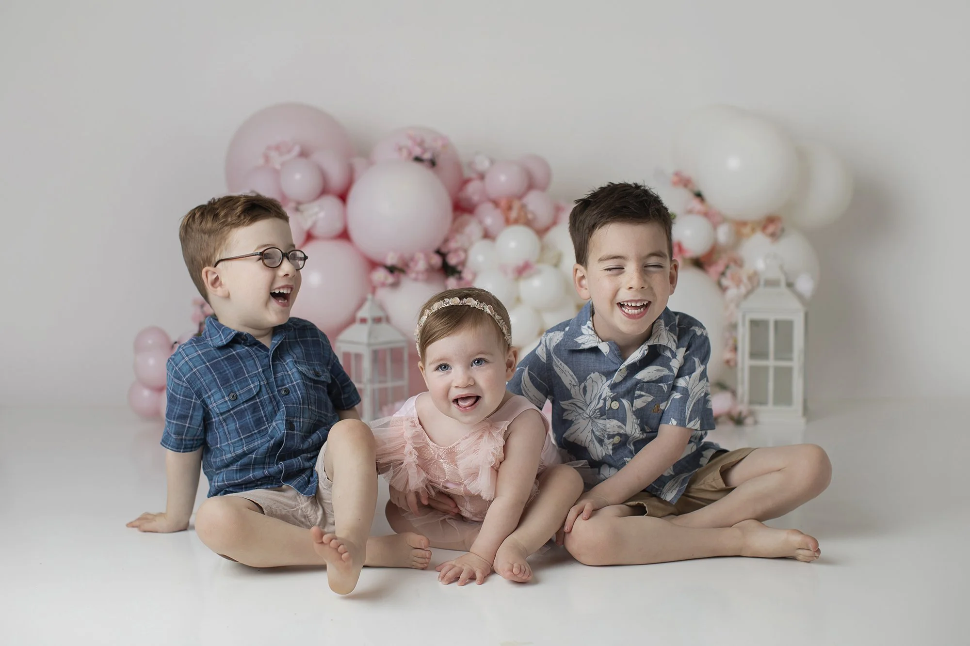 Four children sitting on the floor in front of a balloon and flower backdrop, laughing and smiling.