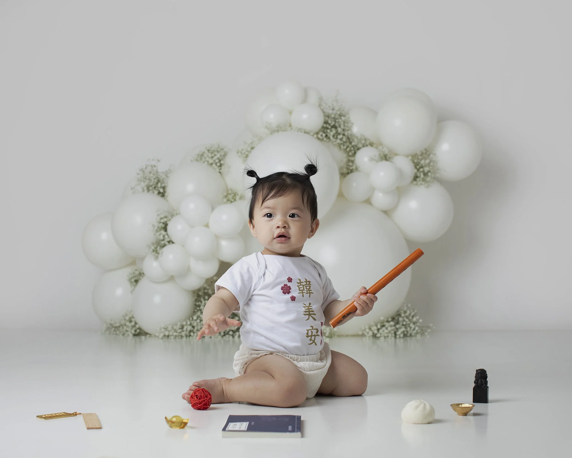 A young child with black hair in pigtails sitting on the floor, holding a long orange object, surrounded by various decorative items including a red ball of string, a black figurine, a small gold bowl, a black notebook, and other small objects. Behin