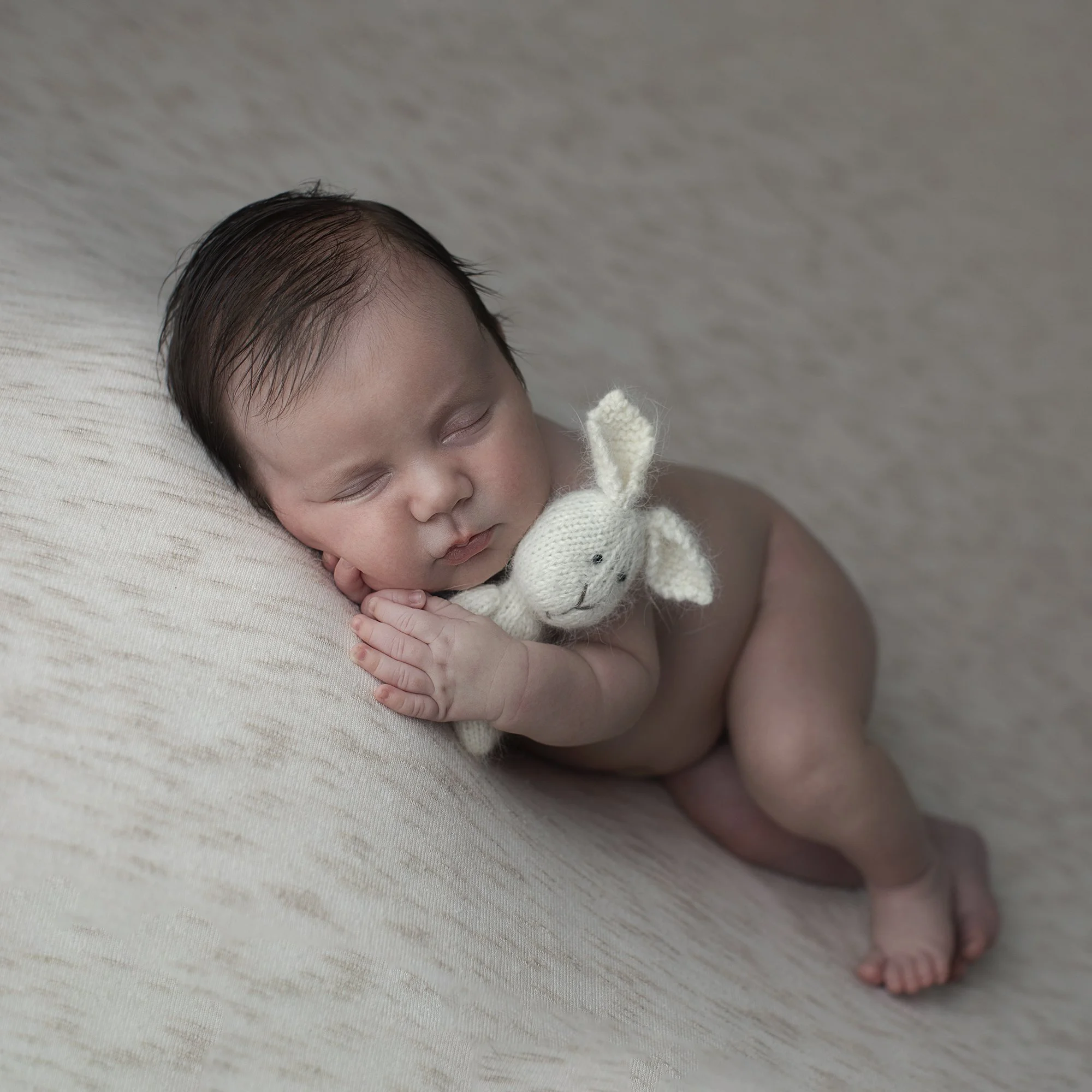 Close-up of a sleeping newborn baby with black hair, lying on a soft surface, wearing a white knitted garment with a small animal design, and resting their hand near their face.