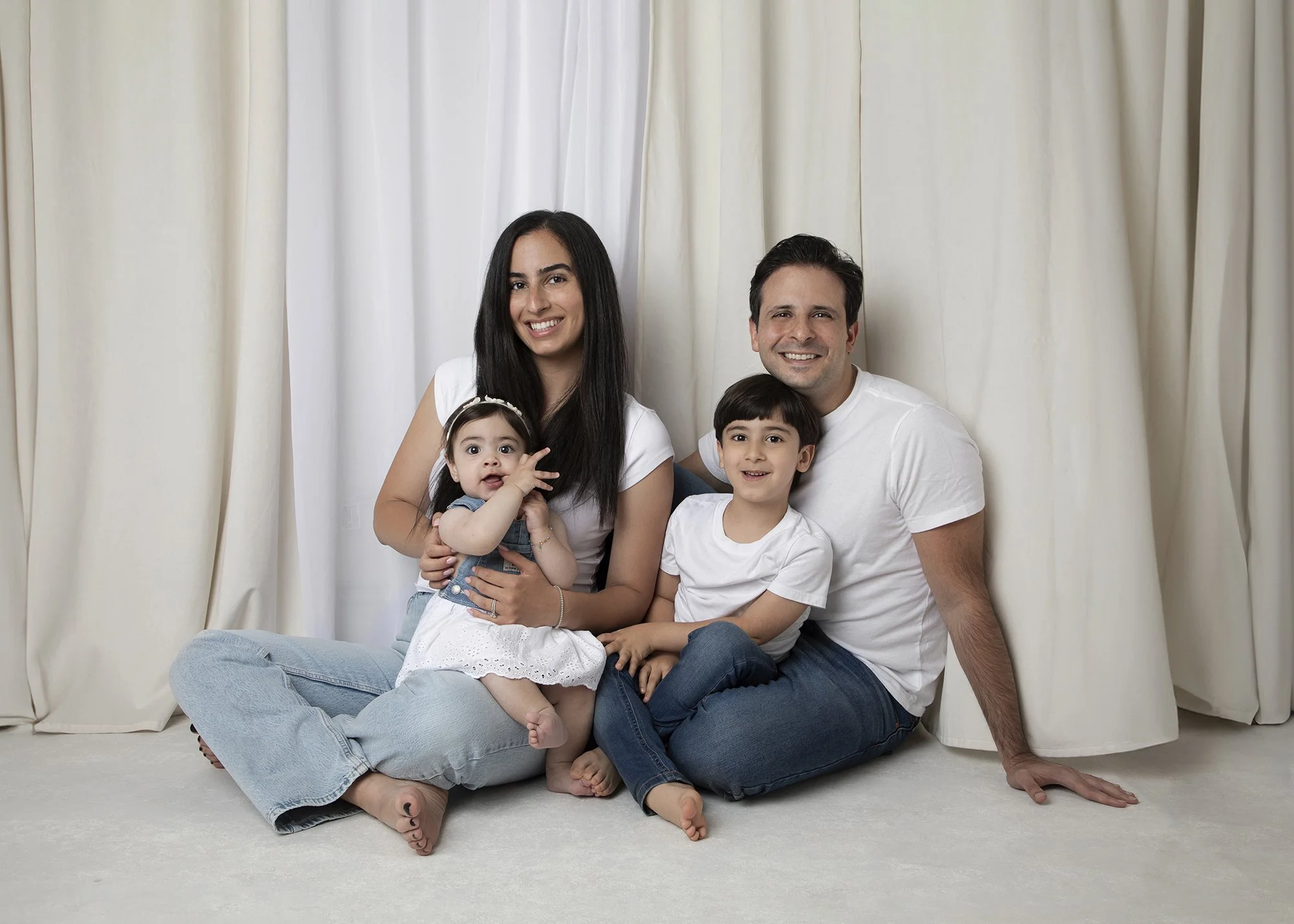 A smiling family of four sitting on the floor in front of cream-colored curtains. The mother has long dark hair, the father has short dark hair, the young girl has long dark hair, and the young boy has short dark hair.