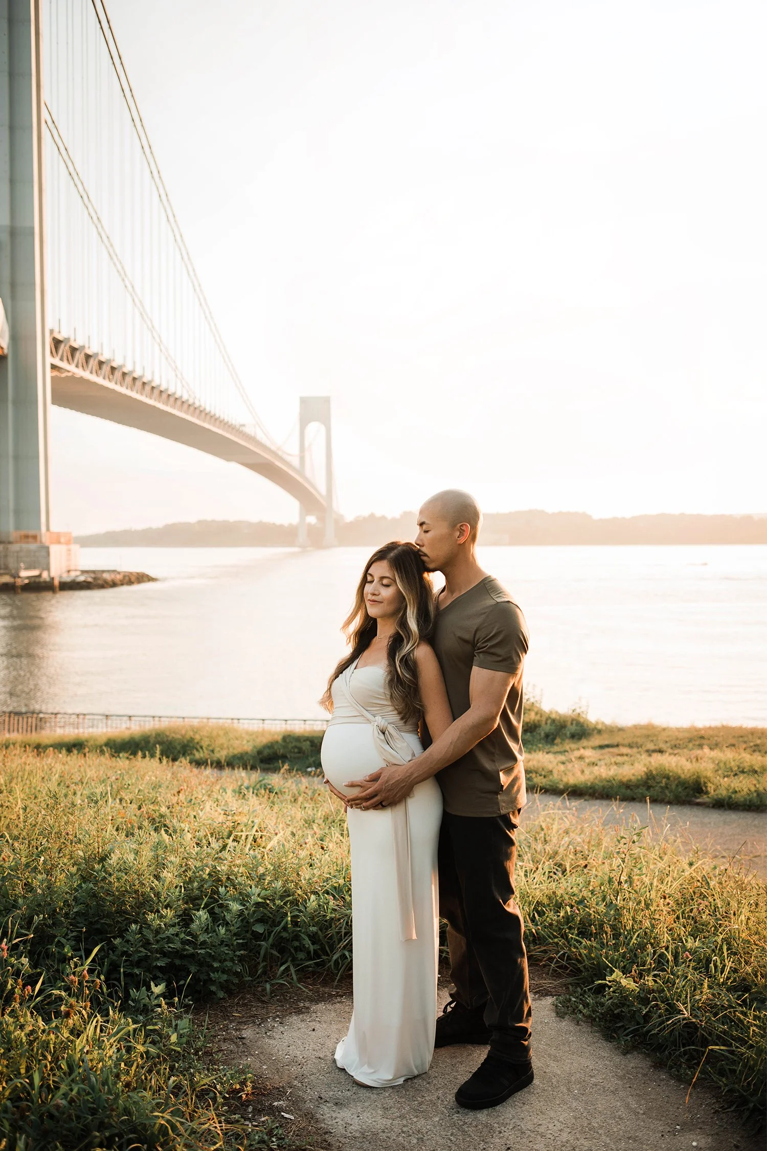 A pregnant woman and a man are standing closely together outdoors near water, with the woman’s hand on her belly and the man’s hand over hers. They are in front of a suspension bridge at sunset, with green plants and grass around them.