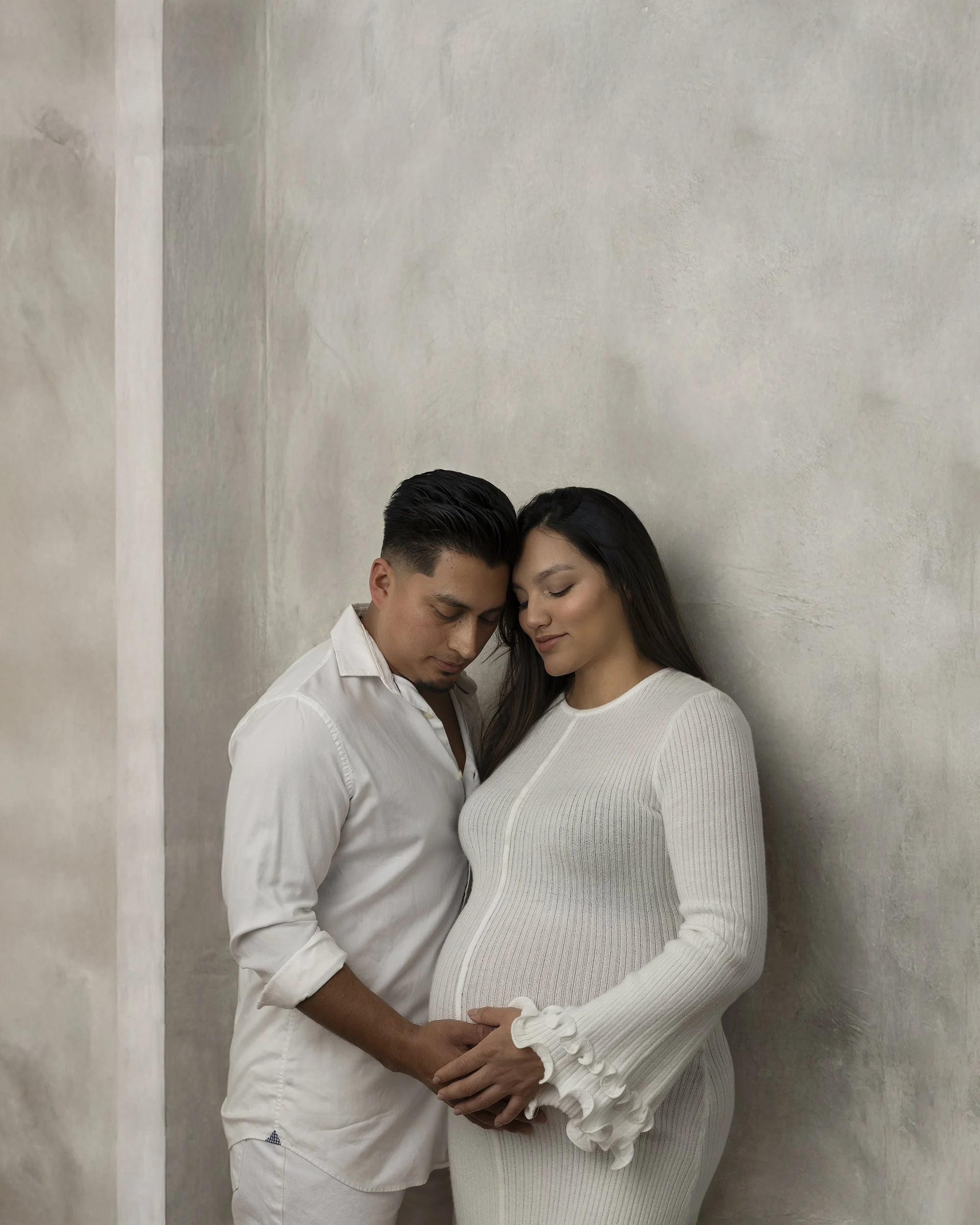 A man and a woman are standing close together, with their foreheads touching and eyes closed, in a loving, intimate gesture. The man has short black hair and is wearing a white shirt. The woman has long black hair and is wearing a white, ribbed, long