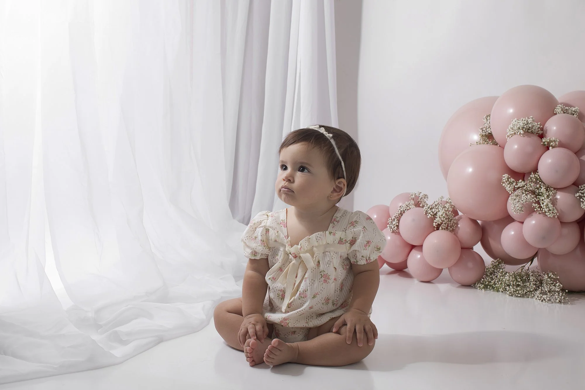 A young girl sitting on the floor in front of a backdrop of pink balloons and white curtains. She has short brown hair with a white headband and wears a floral dress. The girl appears to be looking slightly upward with a neutral expression.