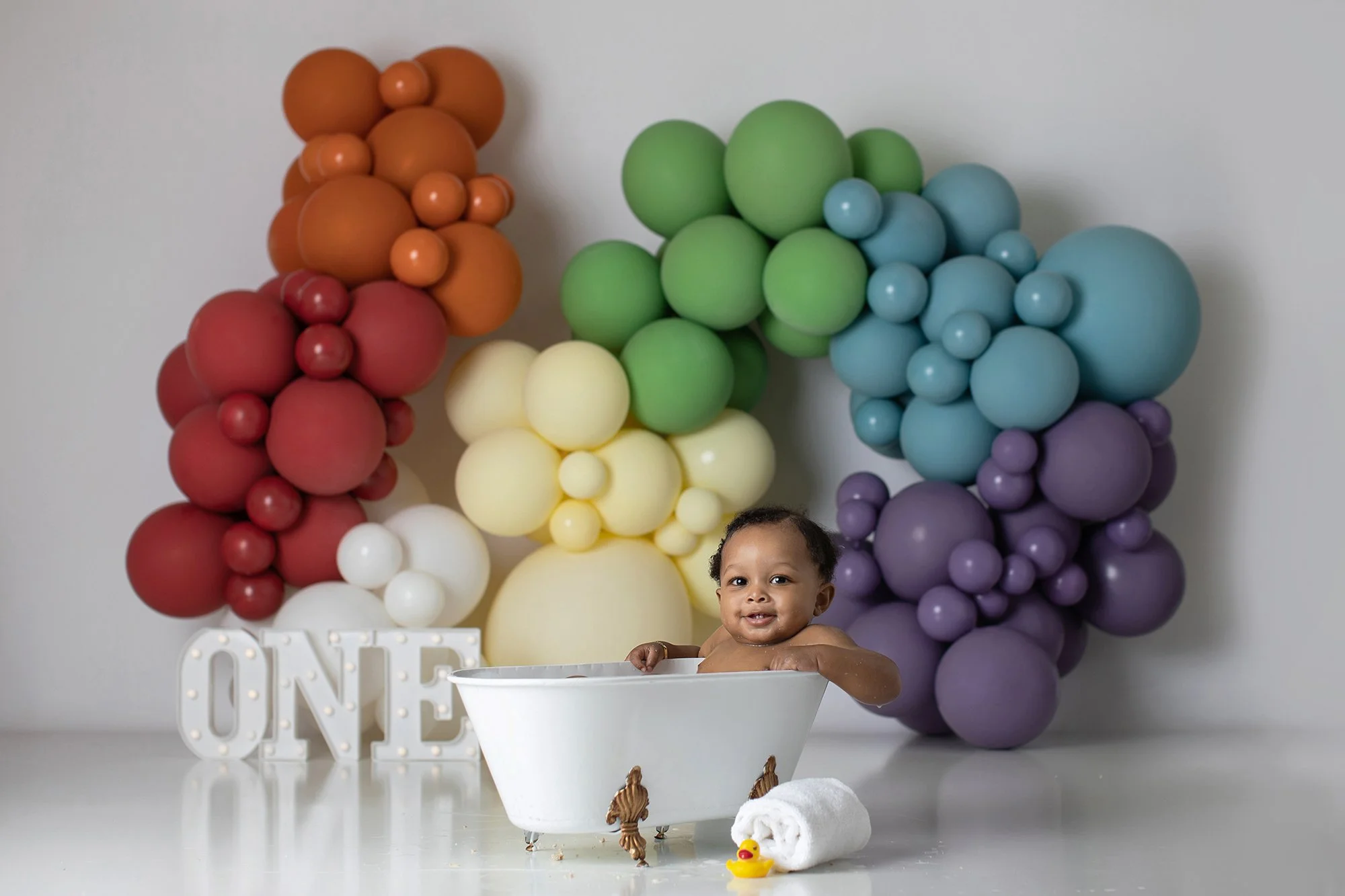 A smiling toddler sitting in a white bucket with a white background decorated with colorful balloons arranged in clusters resembling clouds. There are balloons in colors of red, orange, yellow, green, blue, purple, and white. Part of a white decorati