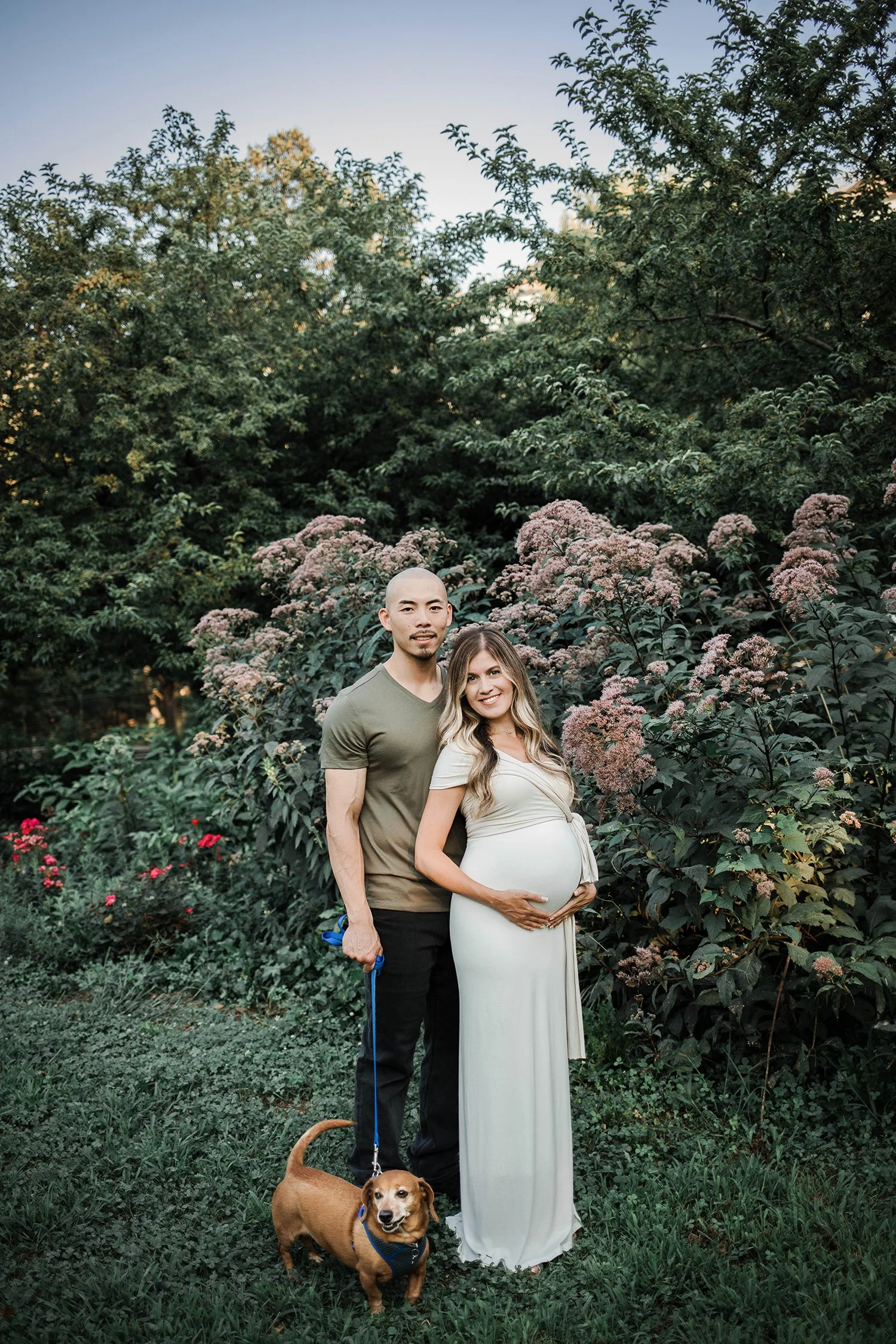 A pregnant woman in a white dress standing outdoors in a garden next to a man holding a dog leash, with a small dog at their feet. They are surrounded by green bushes and trees.