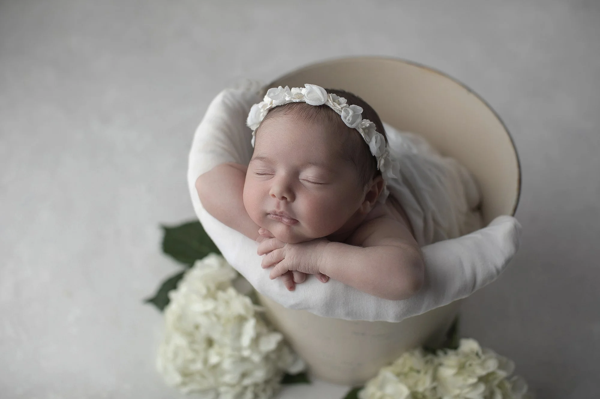 A sleeping newborn baby with a floral headband, resting on hands, surrounded by white flowers.