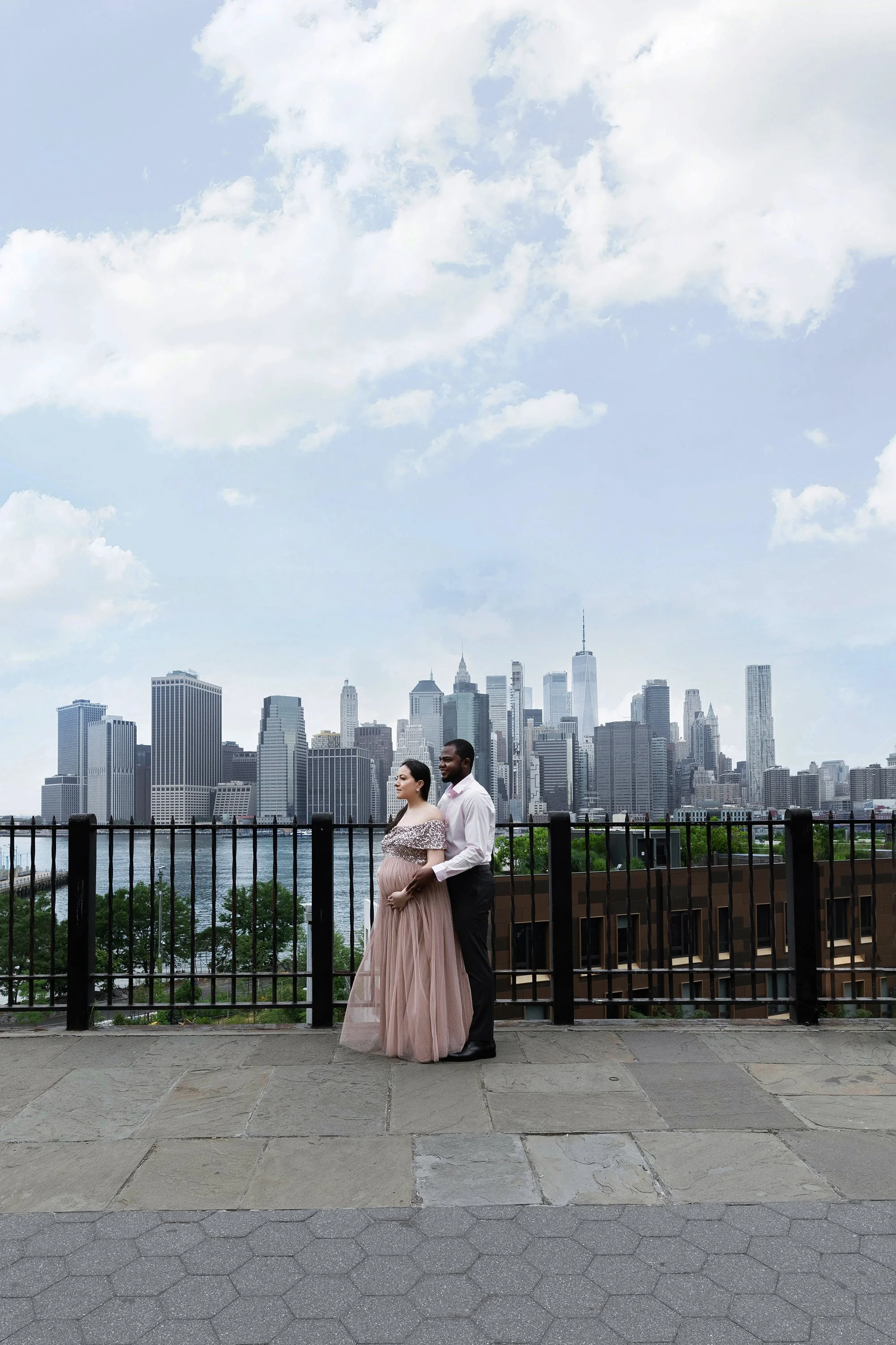 A couple, a pregnant woman in a pink gown and a man in a white shirt and black pants, stand close together by a black fence, with a view of a city skyline and water behind them. The sky is partly cloudy.