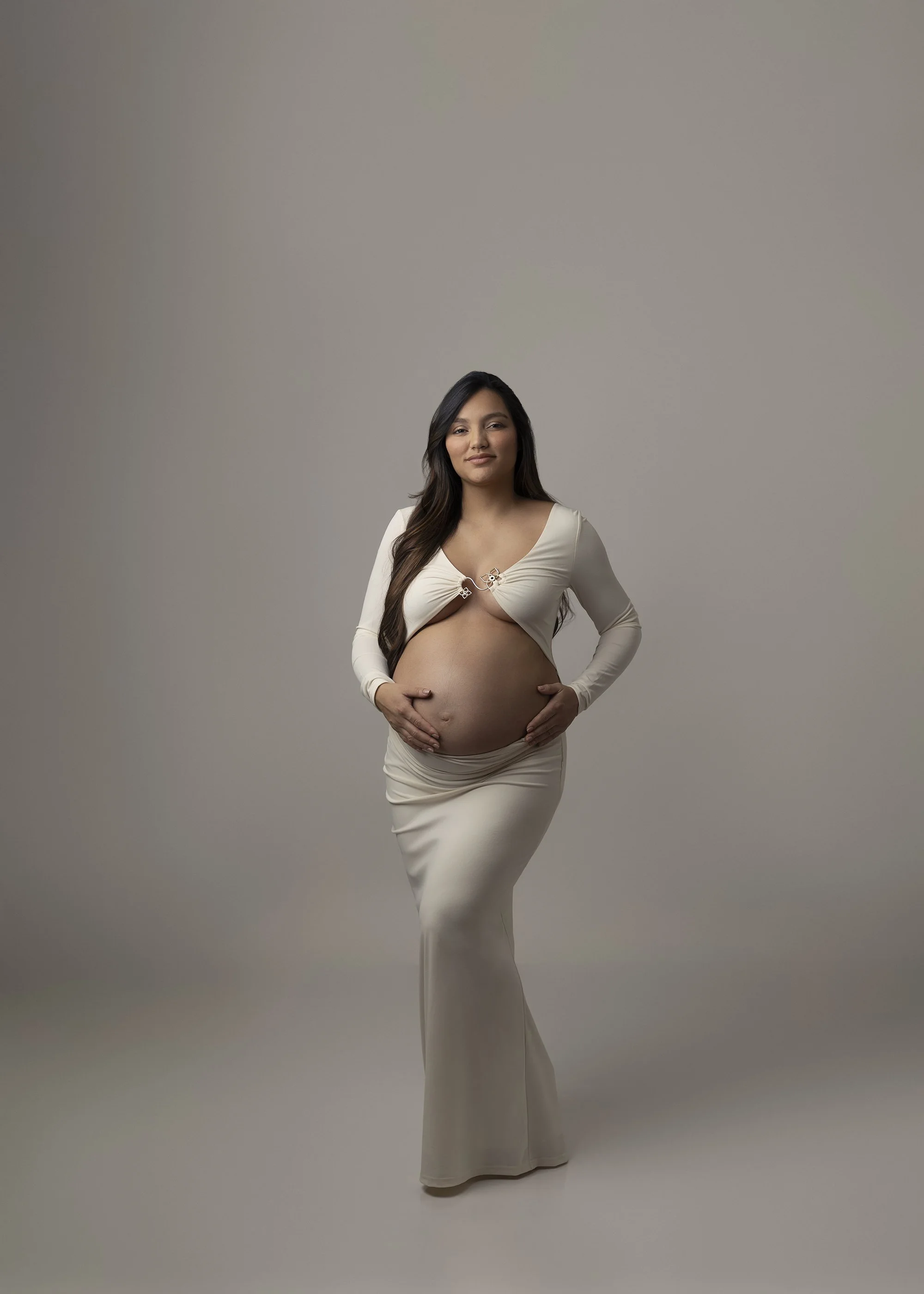 Pregnant woman with long dark hair wearing a white dress with cutouts, standing against a plain gray background.