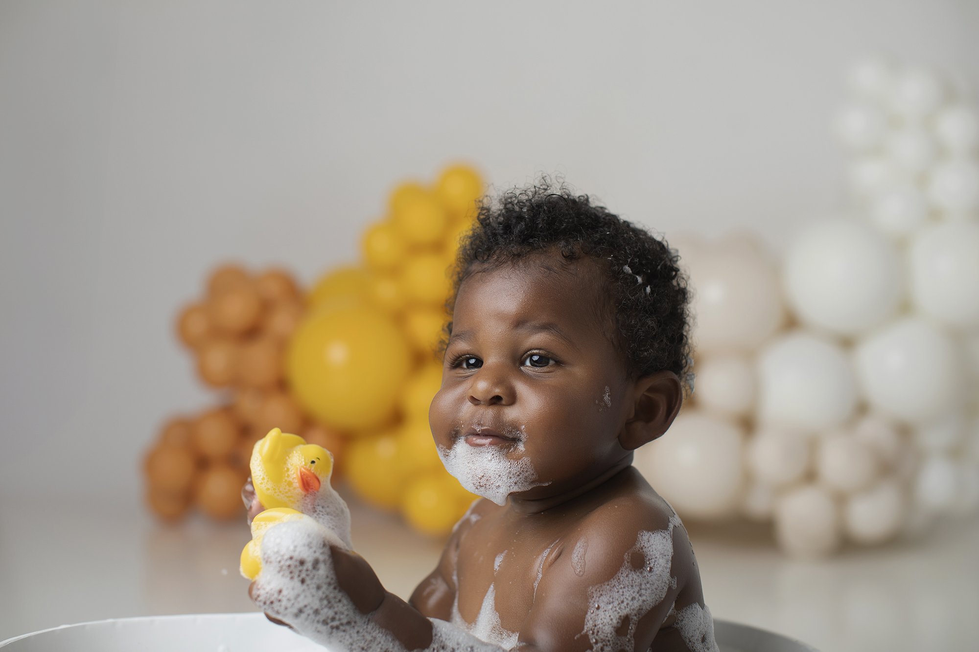 A young child with curly hair taking a bath with soap and bubble foam, holding a yellow rubber duck in a playful setting with colorful balloons in the background.