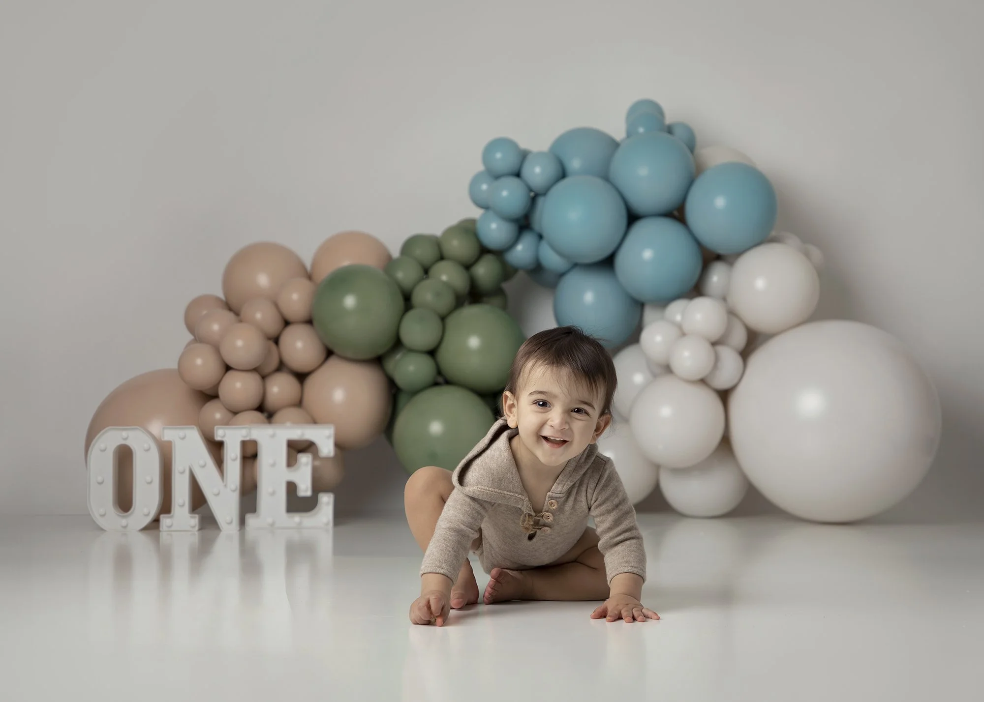 A young boy crawling on the floor in front of a backdrop of large pastel-colored balloons and a decorative sign spelling "ONE" for a first birthday celebration.