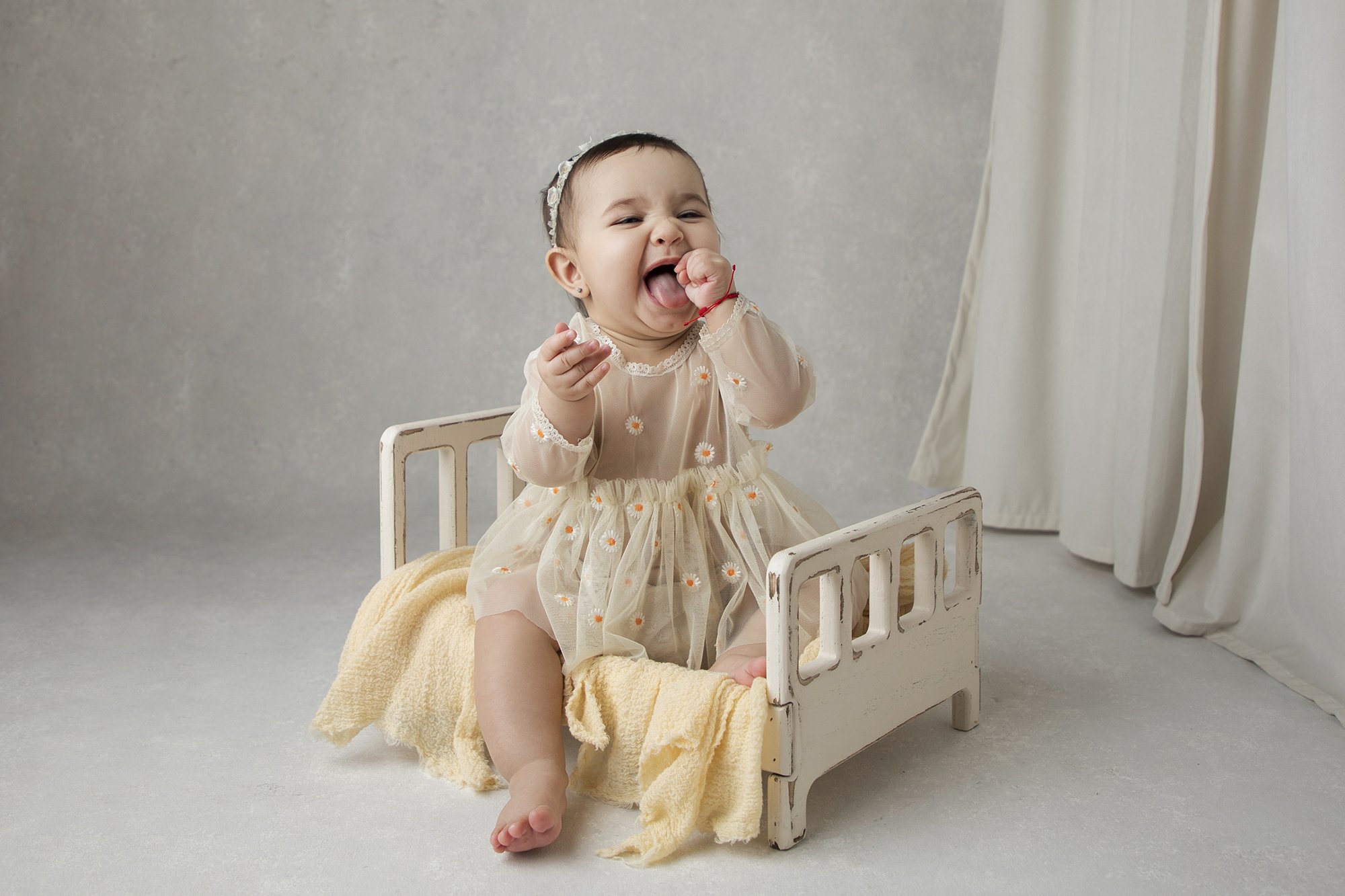 A baby girl with short dark hair wearing a sheer cream dress with daisies, sitting in a small white wooden bed with a yellow blanket, with a neutral background and white curtains.