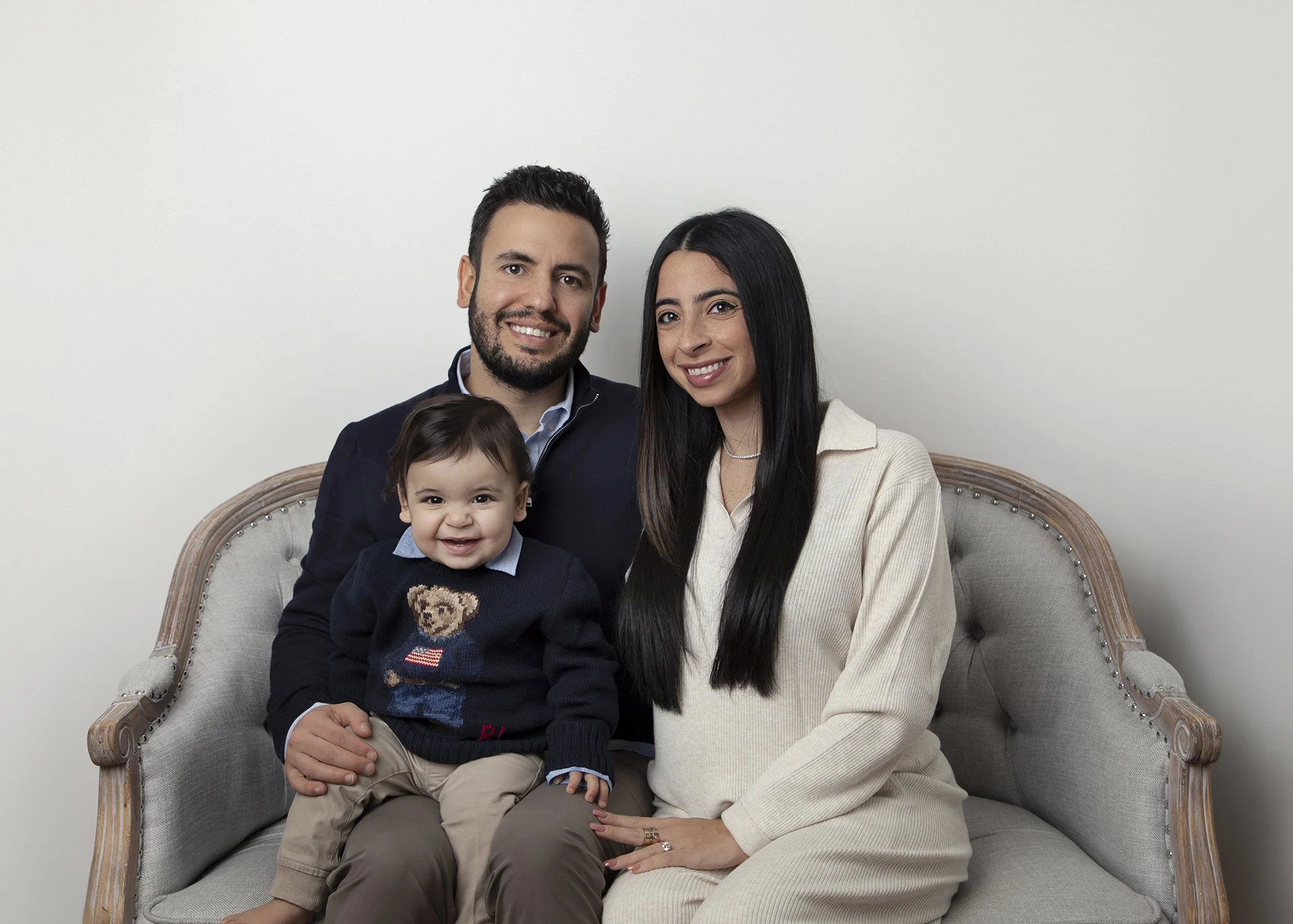 A family of four sitting on a vintage-style sofa against a plain white wall. The family includes a bearded man with dark hair, a woman with long black hair, a young boy with dark hair, and a toddler boy with light brown hair wearing a sweater with a 