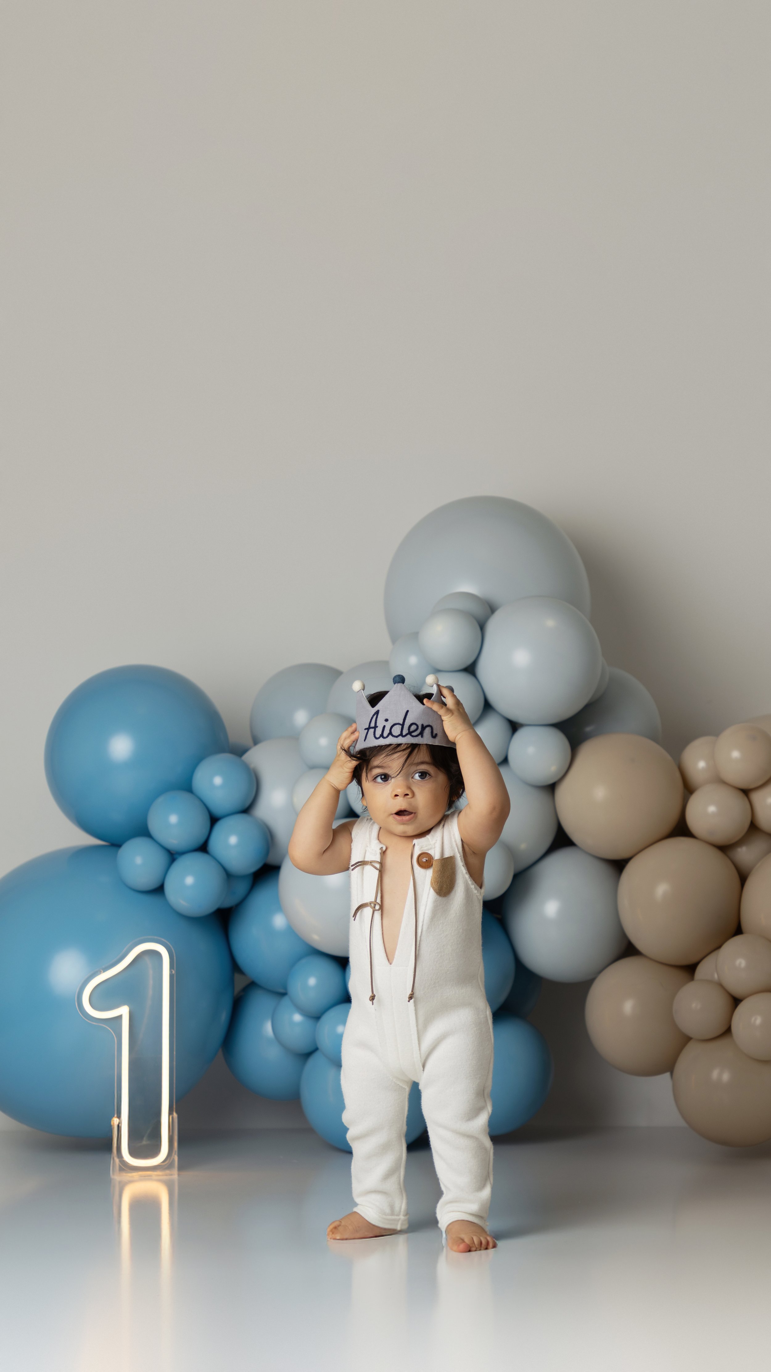 A young child wearing a white jumpsuit holding a gray hat with the name 'Aiden' written on it, standing in front of a backdrop of blue and beige balloons, with a neon number 1 and a lighted number 1 nearby, indicating a first birthday celebration.