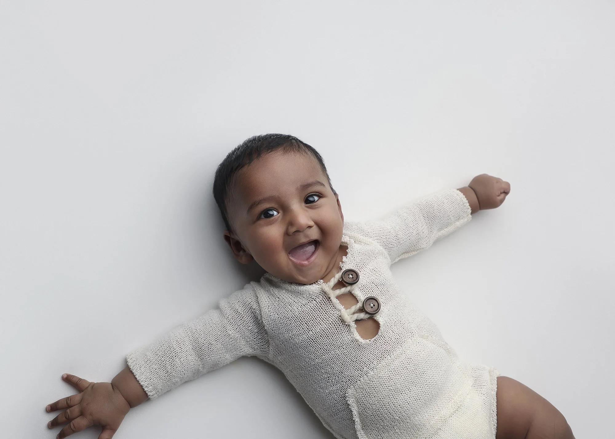 A happy baby lying face up on a white surface, looking up, with arms outstretched and wearing a cream-colored knitted sweater with buttons.