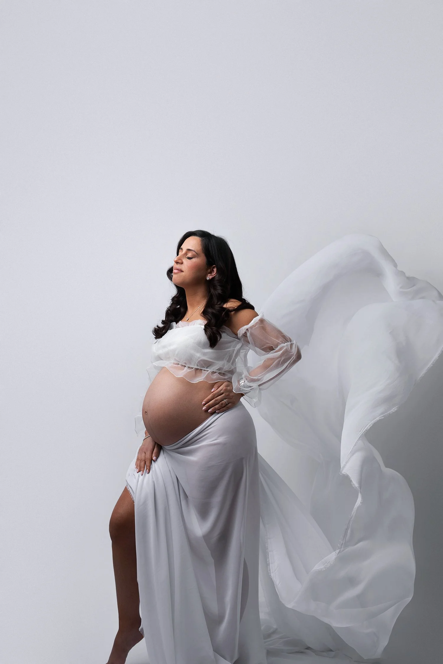 Pregnant woman with long dark hair wearing a white dress and standing against a plain white background, with flowing fabric around her.