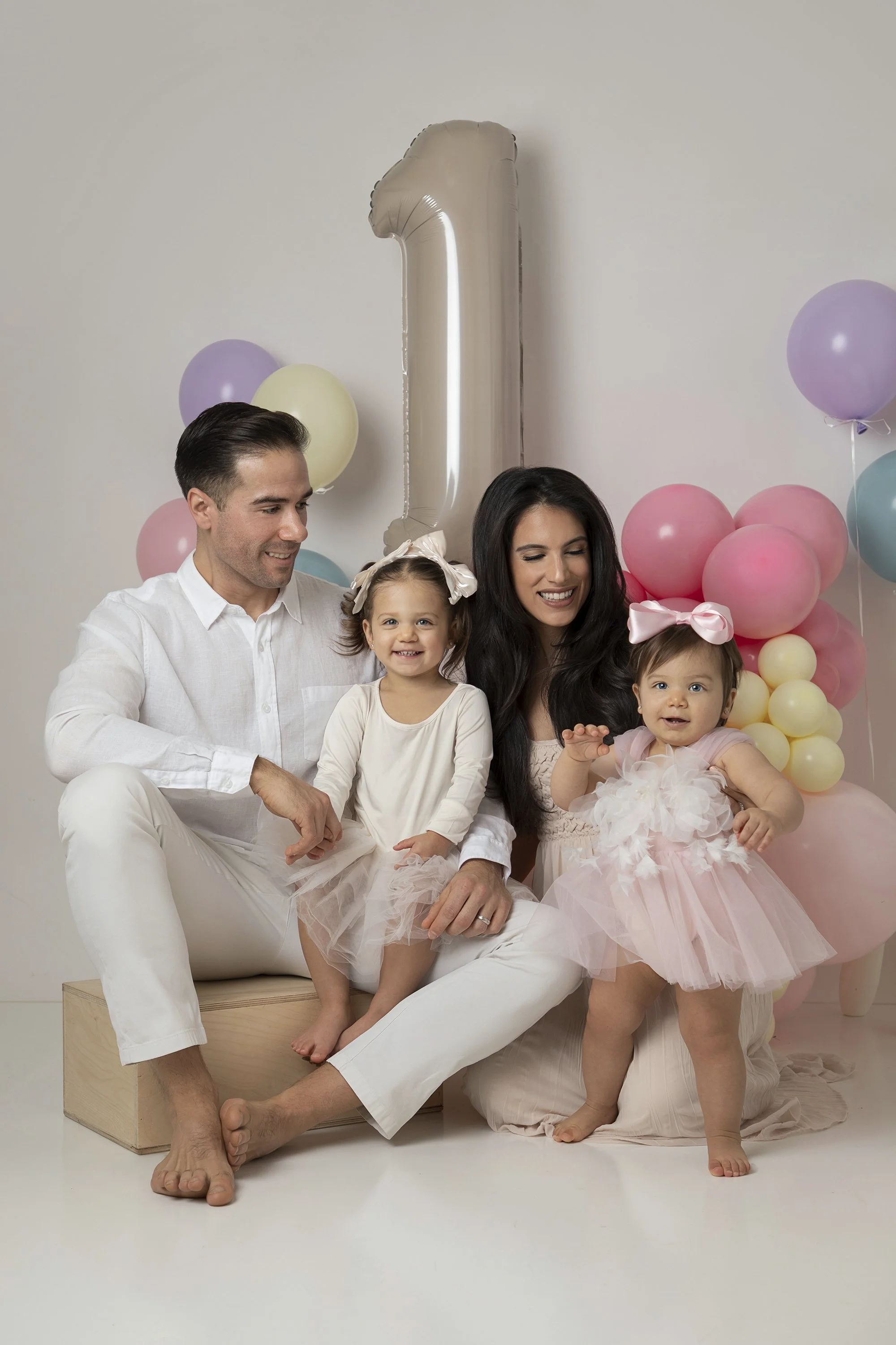 A family of four with two young girls celebrating a birthday, with pastel balloons and a metallic number one balloon in the background.