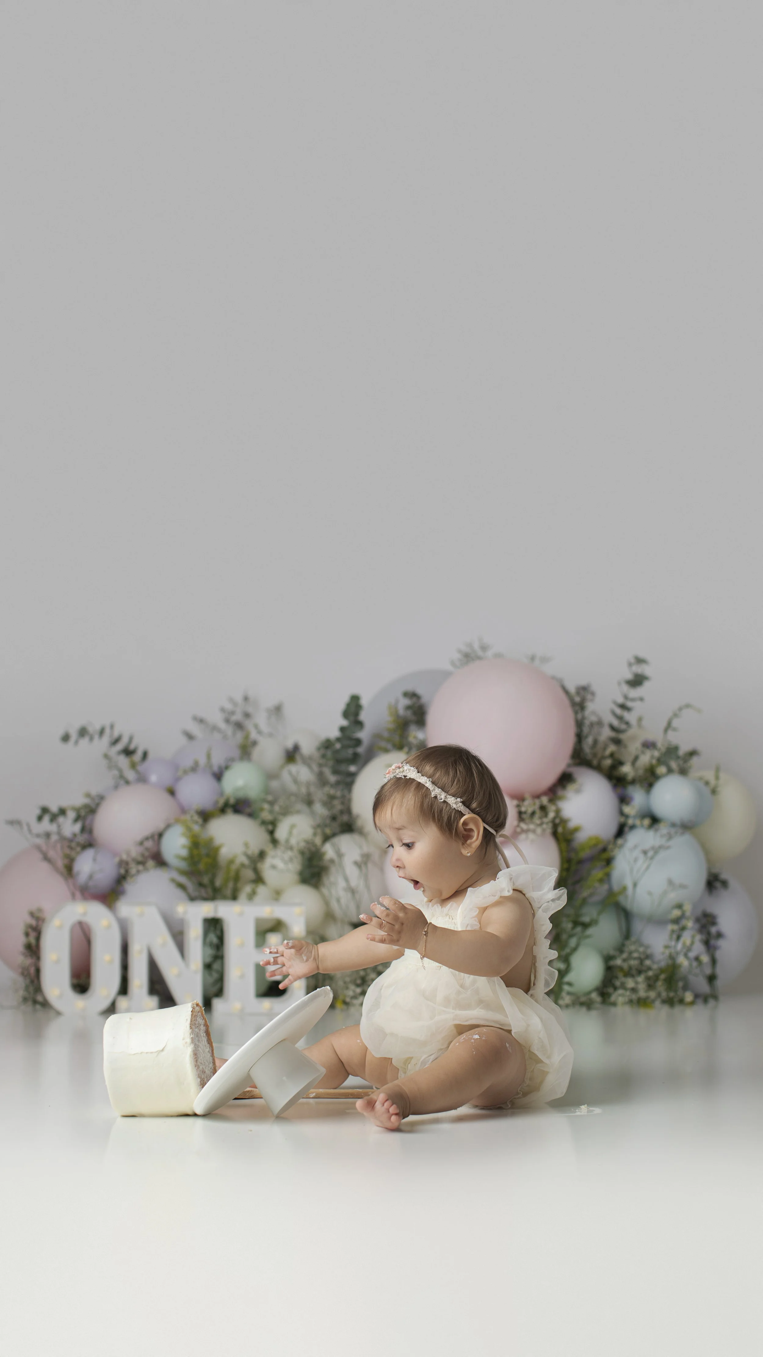 A young girl in a cream dress and headband playing with a cake and toppers at a birthday celebration with pastel balloons and a decorative sign saying 'ONE' in the background.