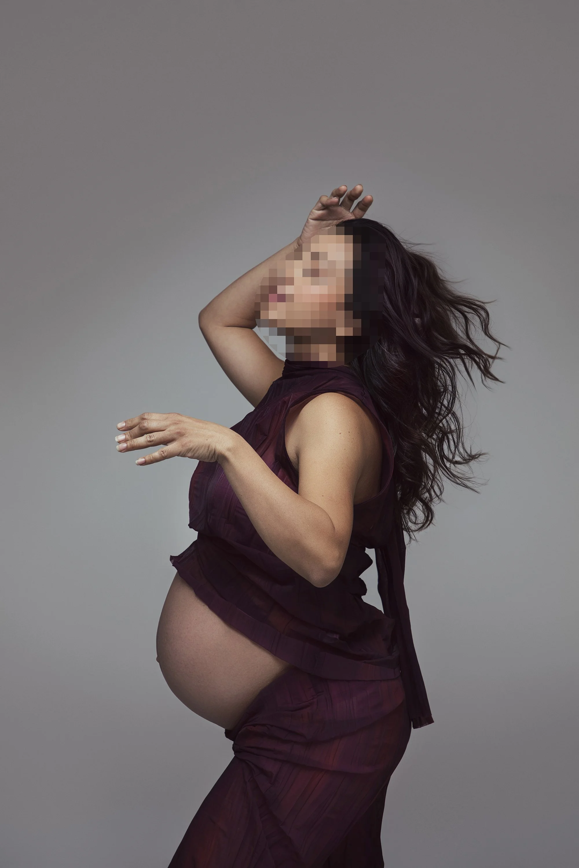 A woman in sheer, dark purple, ruffled dress, with long wavy hair, posing against a plain gray background.