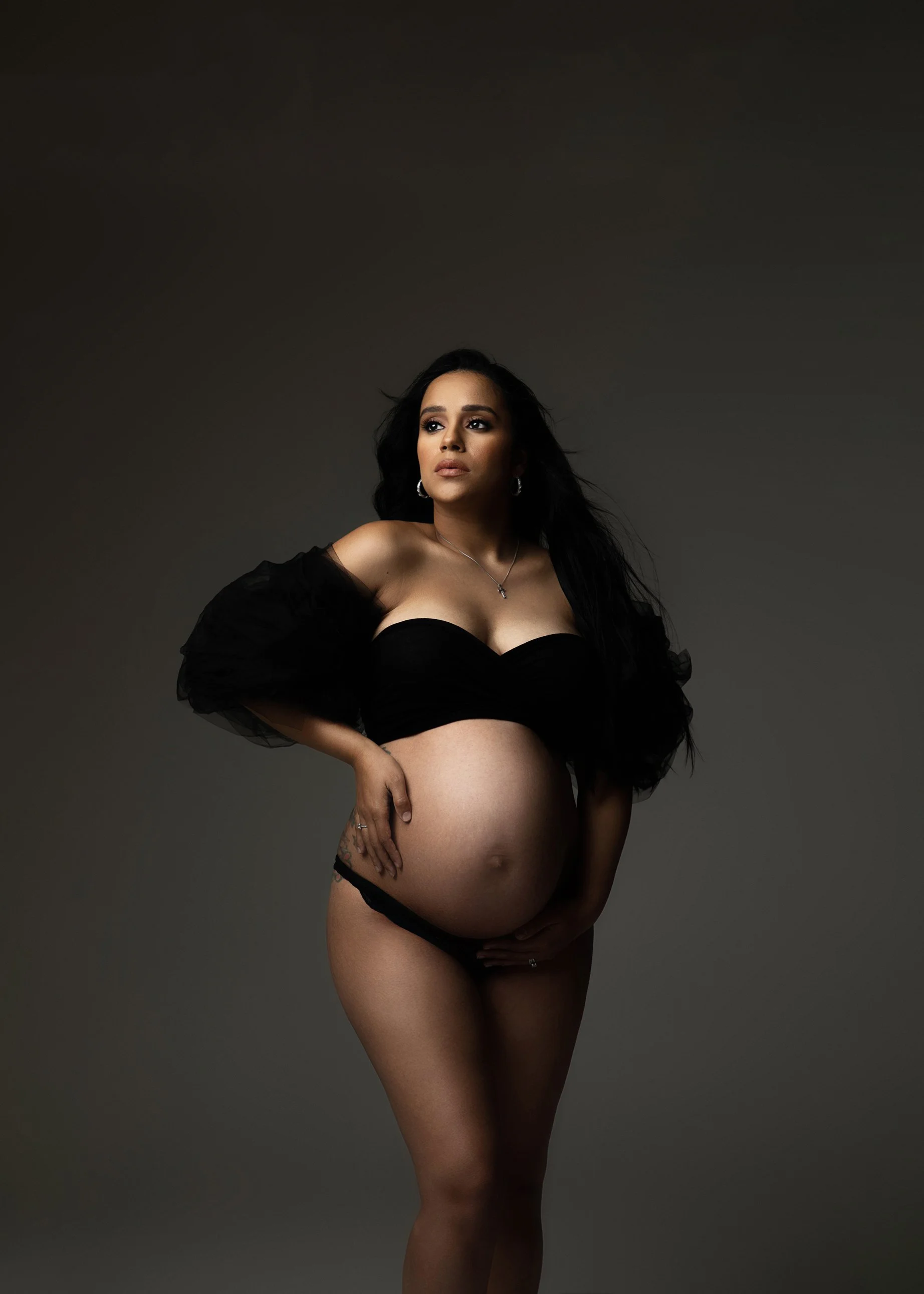 A pregnant woman with dark hair and makeup, wearing a black off-shoulder top with puffed sleeves and black lingerie, standing against a dark gray studio background. She has jewelry, including earrings and a necklace, and is posing with one hand on he