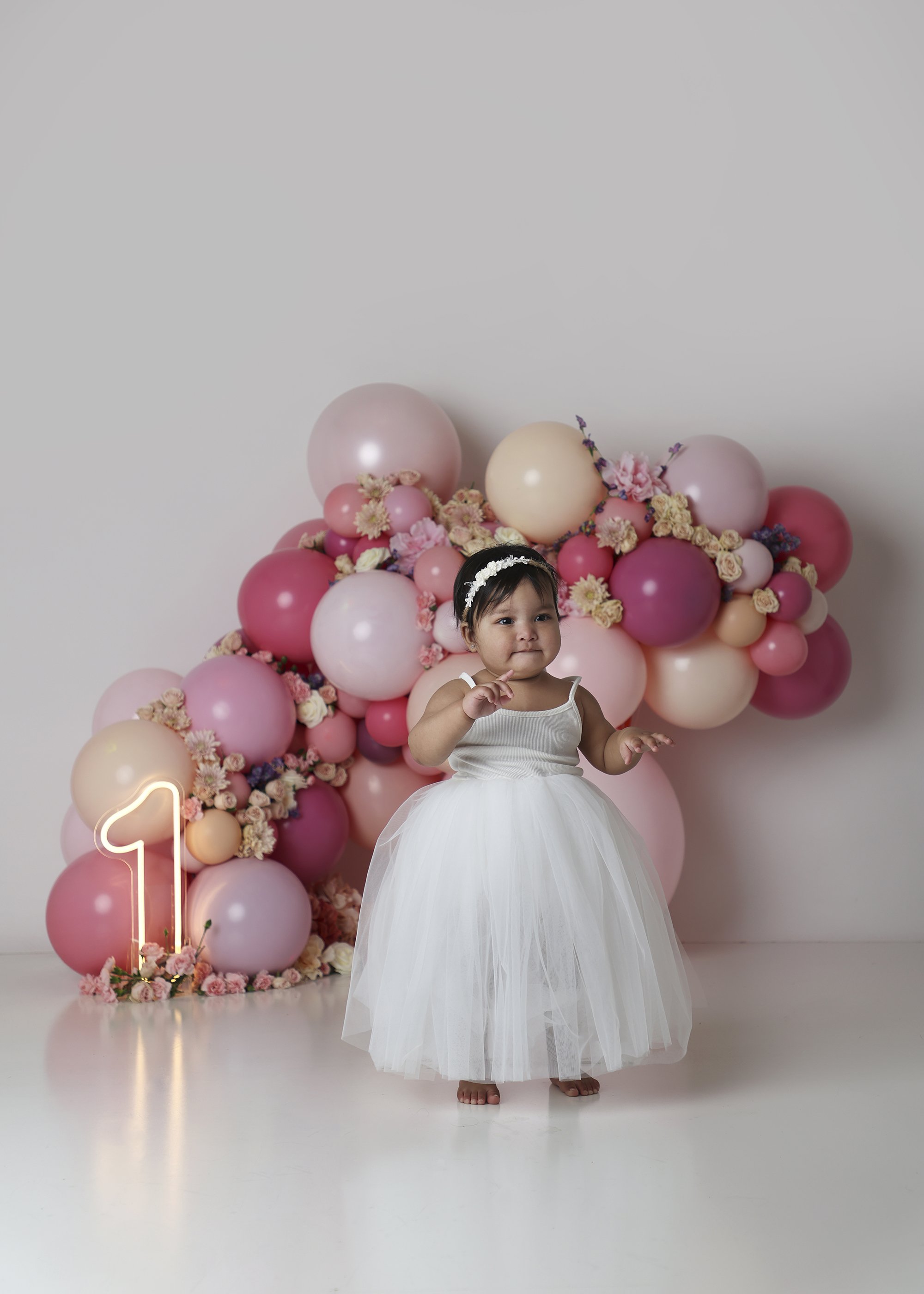 A young girl in a white dress and floral headband standing in front of a pink balloon and flower backdrop, with a neon number one sign to her left.