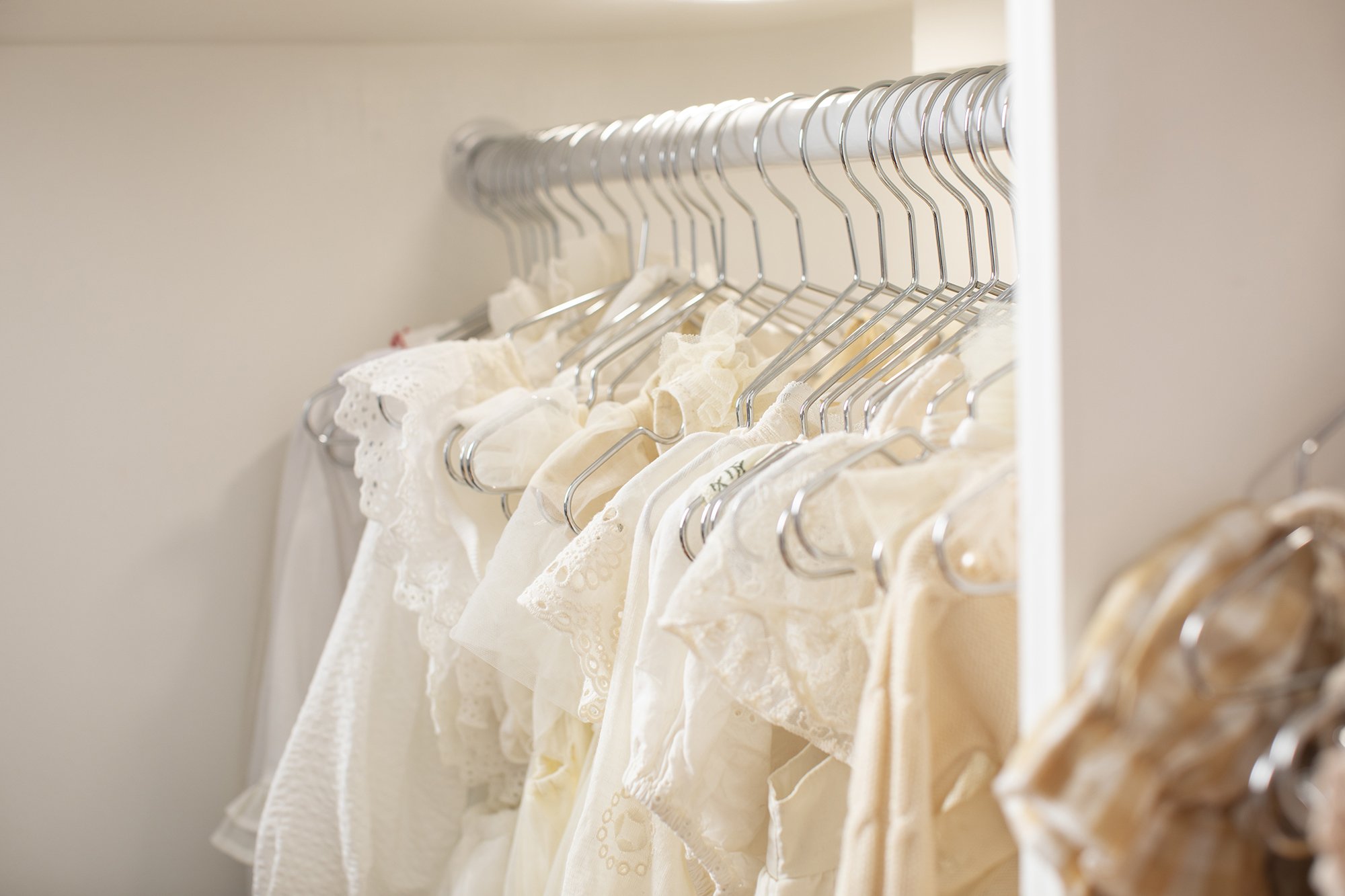 A closet with a row of white and beige lace and embroidered dresses hanging on metal hangers.