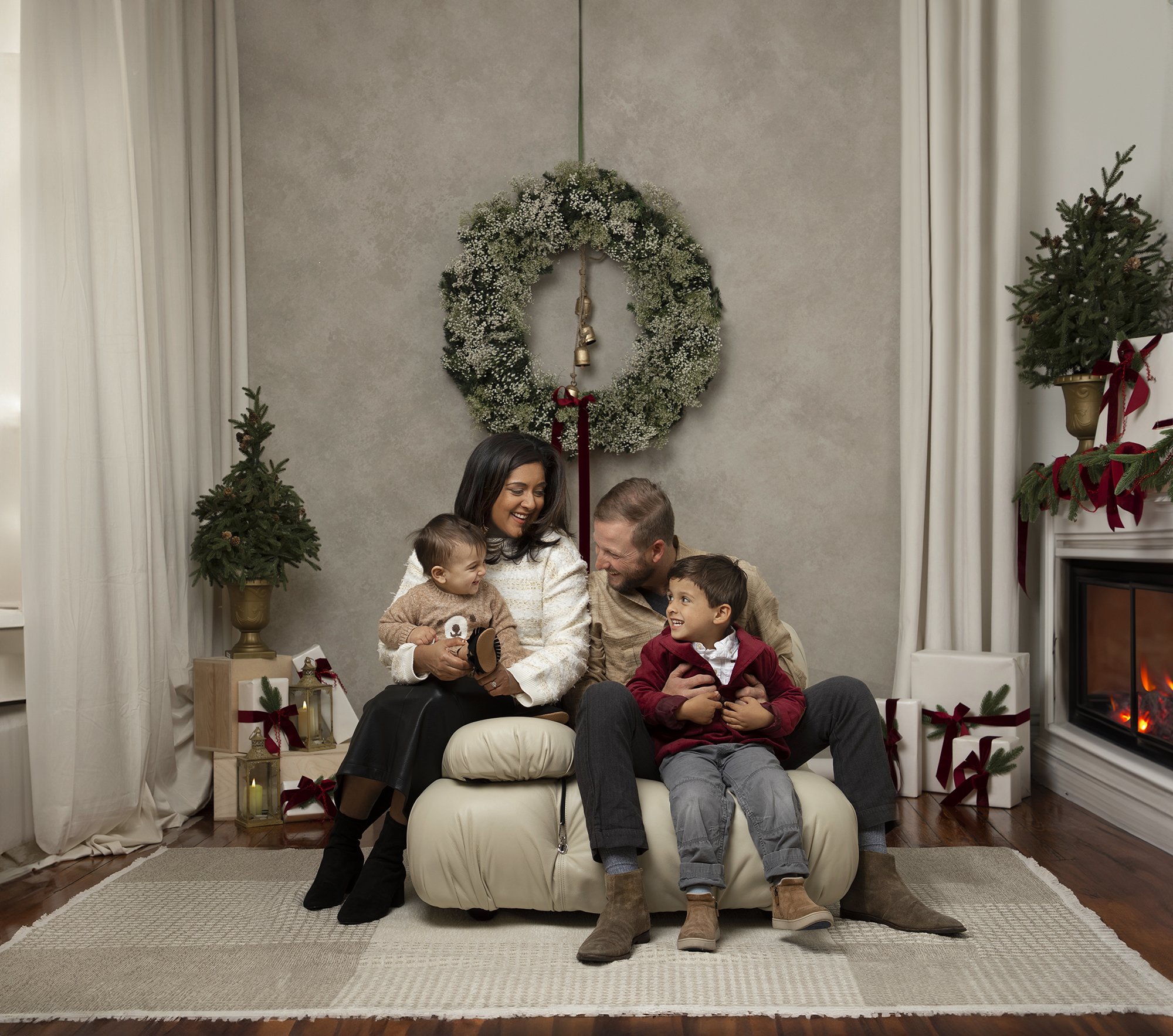 A family of five enjoying Christmas together in a decorated living room with a wreath on the wall, Christmas trees, and wrapped presents. They are sitting on a couch, smiling and interacting with each other.
